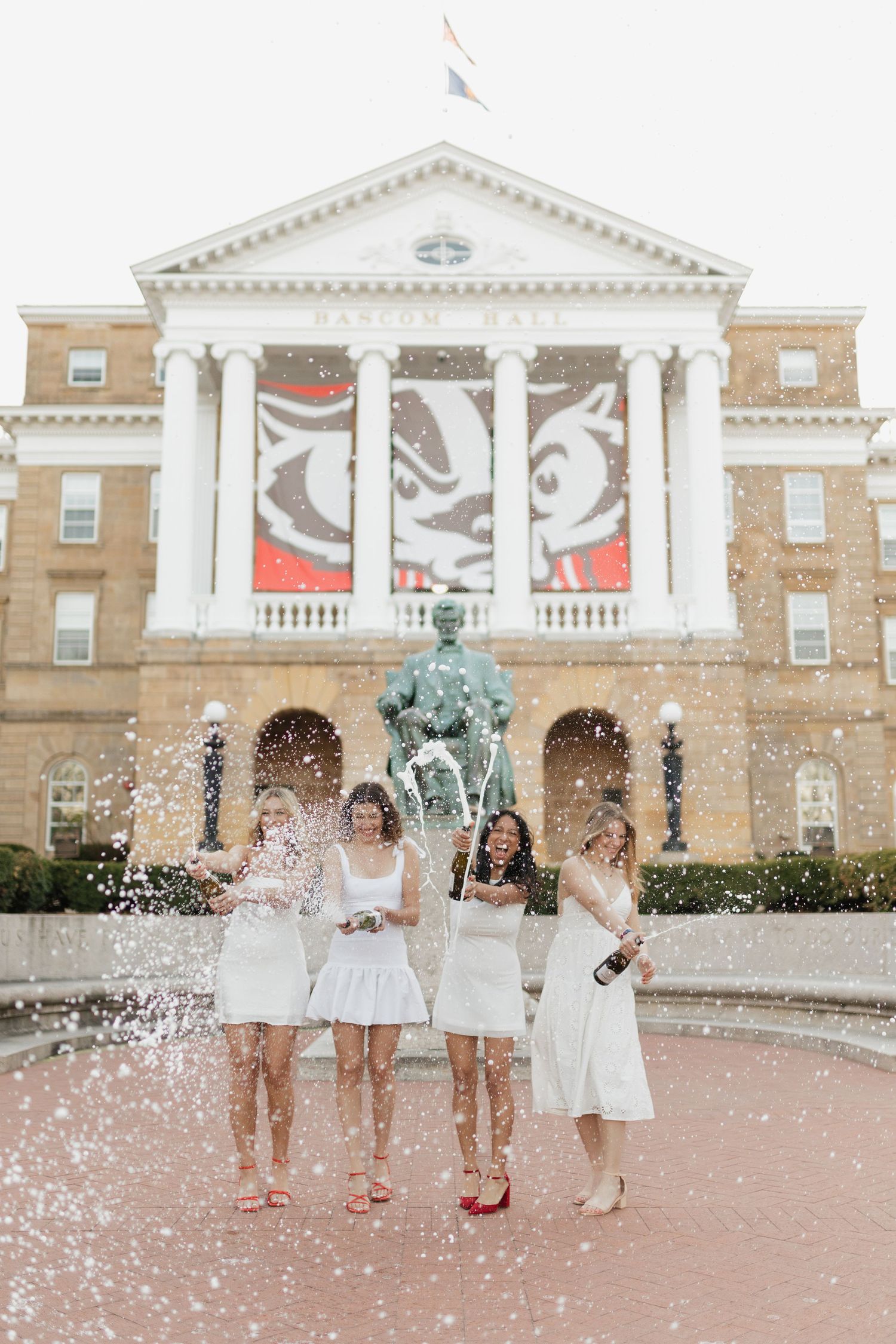 Graduates in white dresses celebrate with champagne spray in front of classical building.
