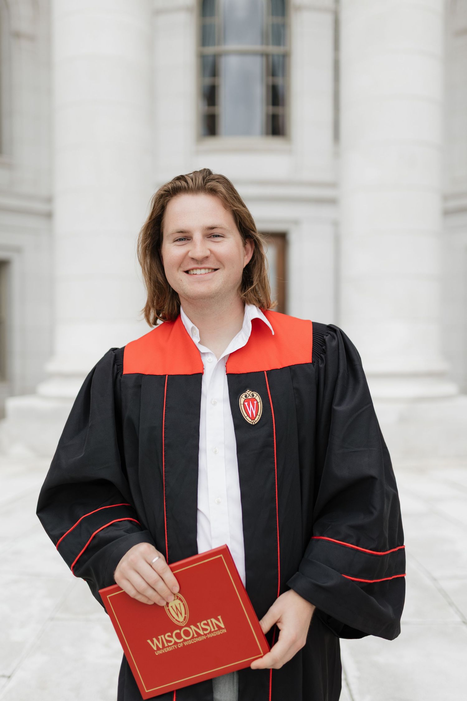 Graduate in black and red graduation gown holds Wisconsin diploma in front of white columns.