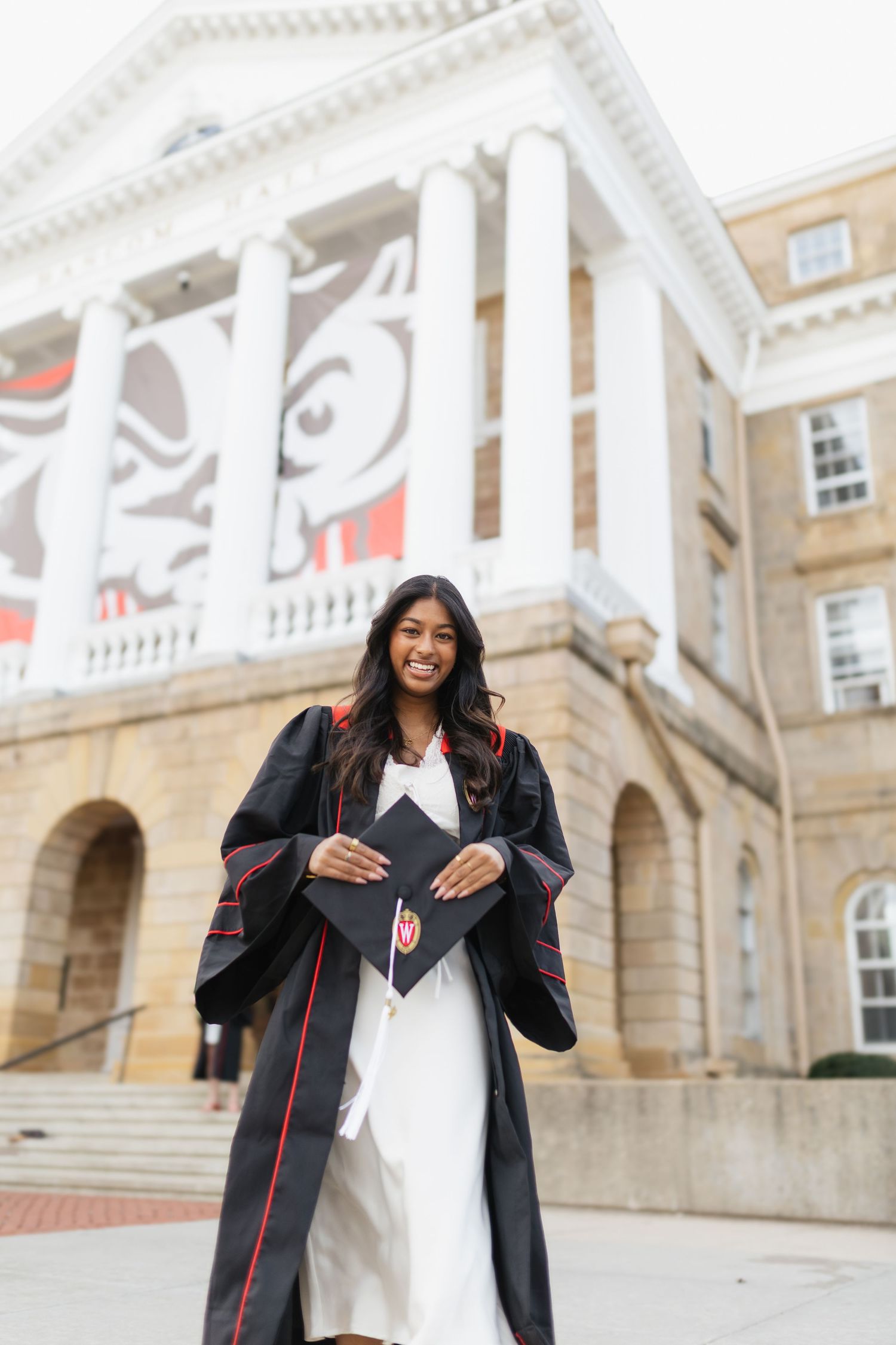 Graduate in cap and gown poses with diploma in front of collegiate building with decorative facade.