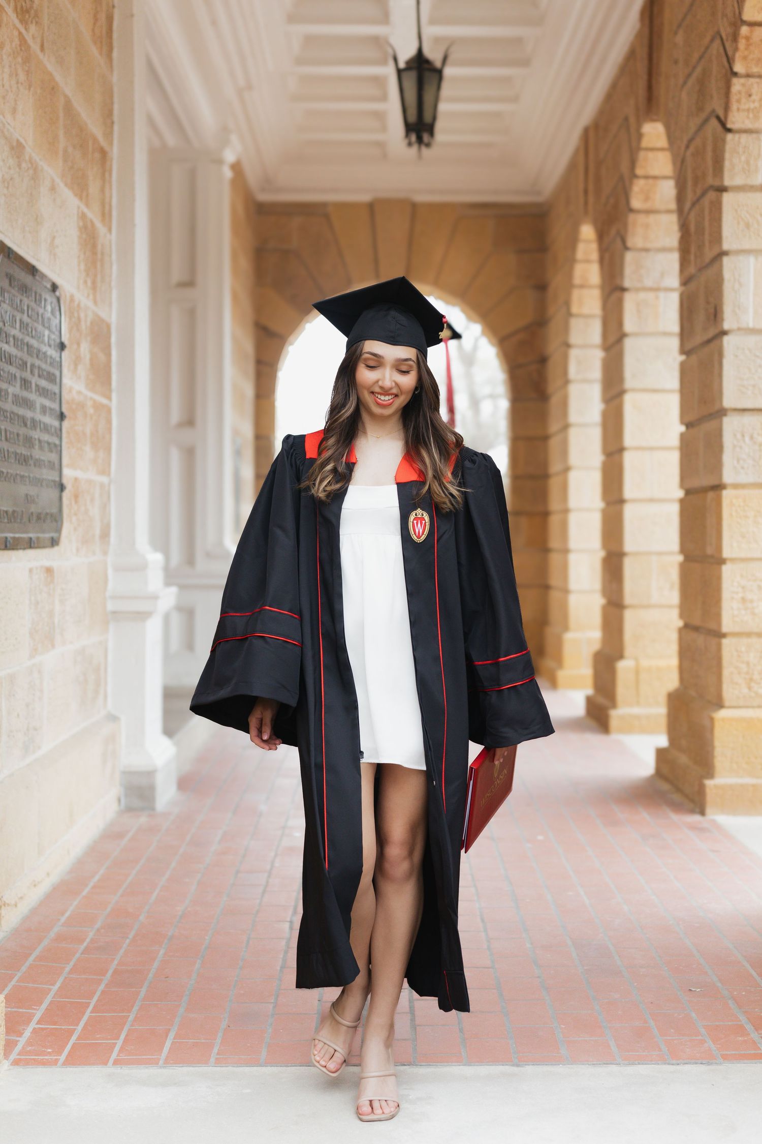 Graduate in cap and gown walks through sunlit stone archway.