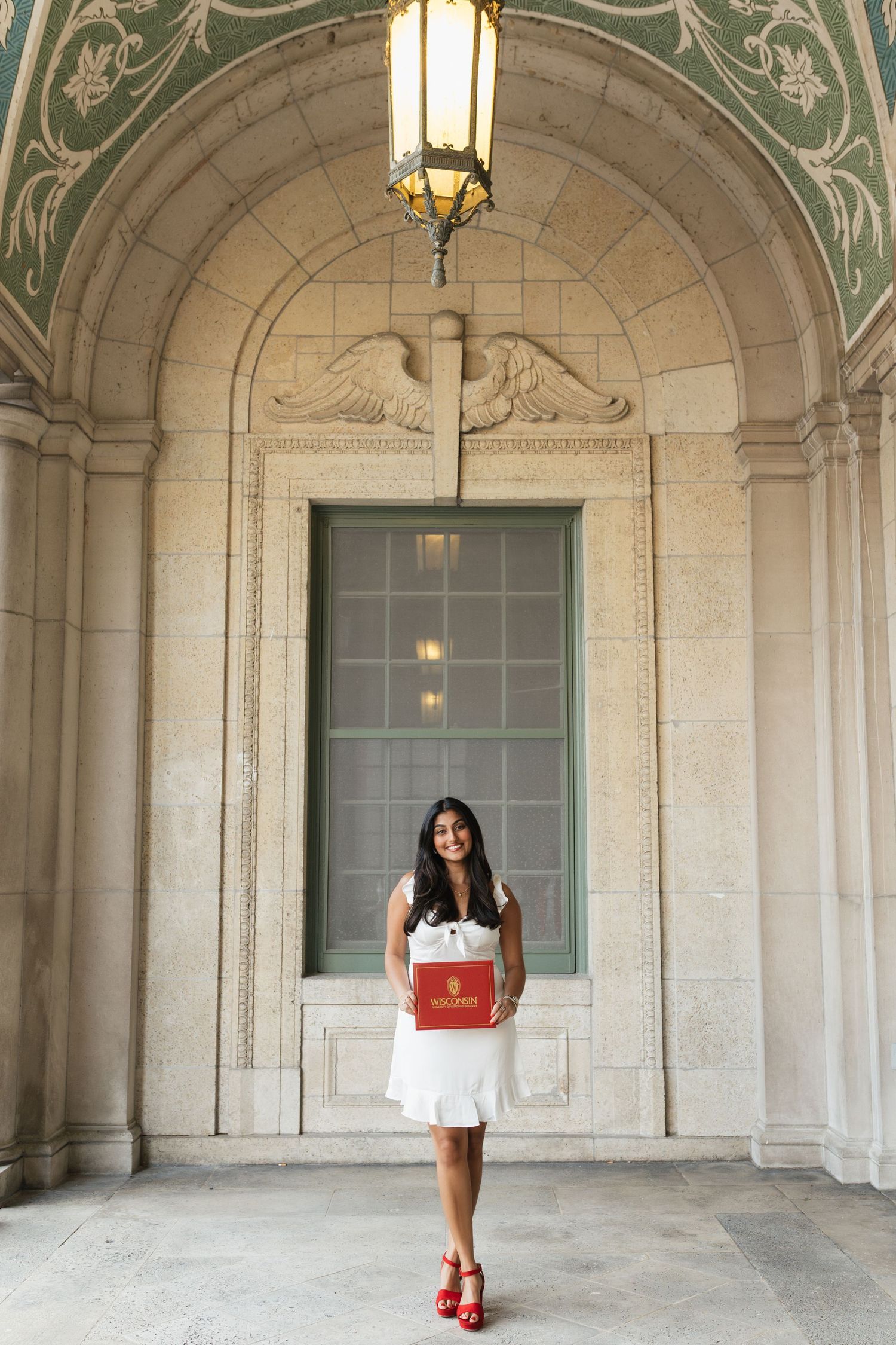 Graduate in white dress and red shoes poses under ornate stone archway with decorative lantern.