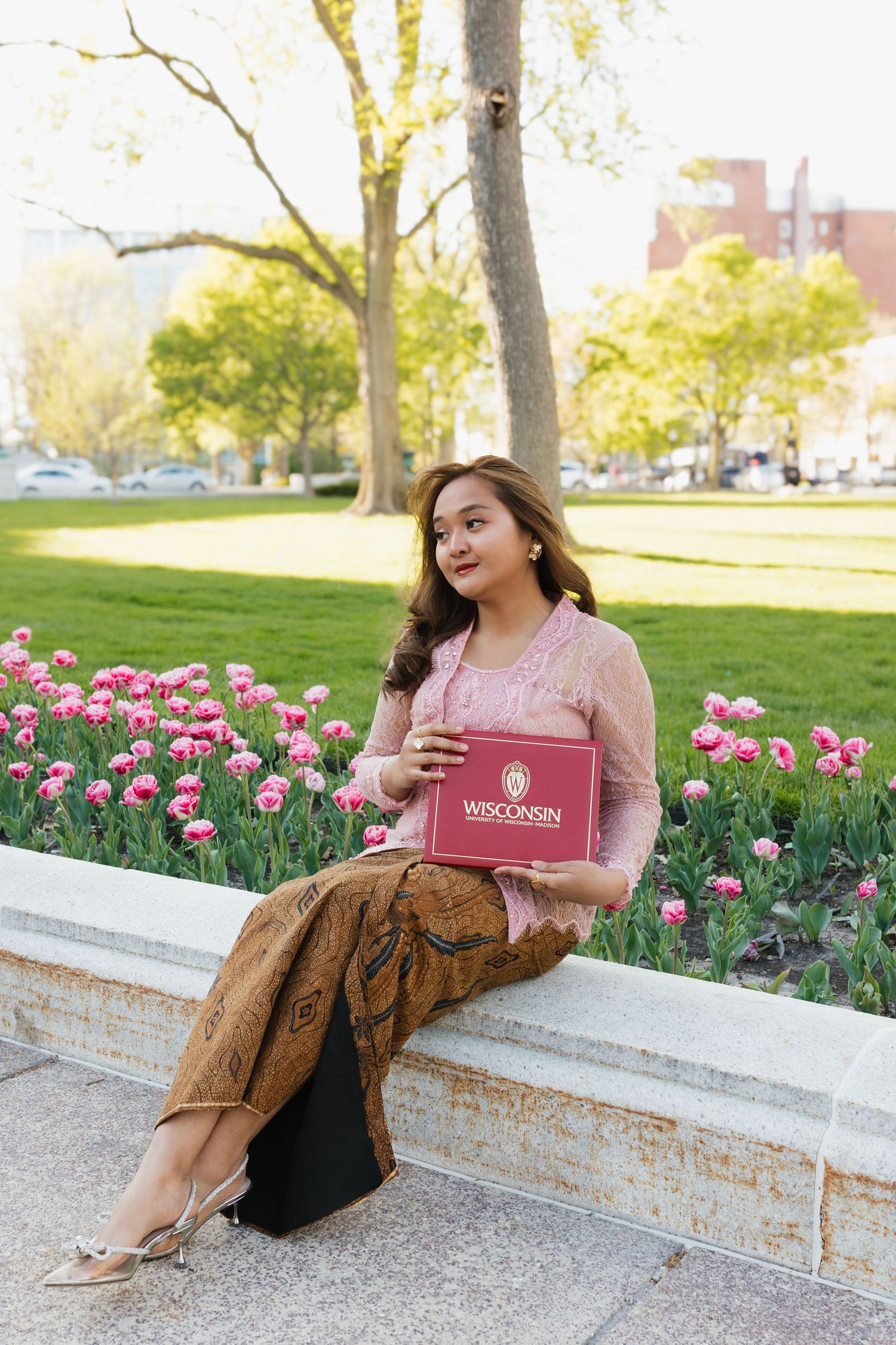 Graduate in pink outfit sitting by tulip garden with university diploma.