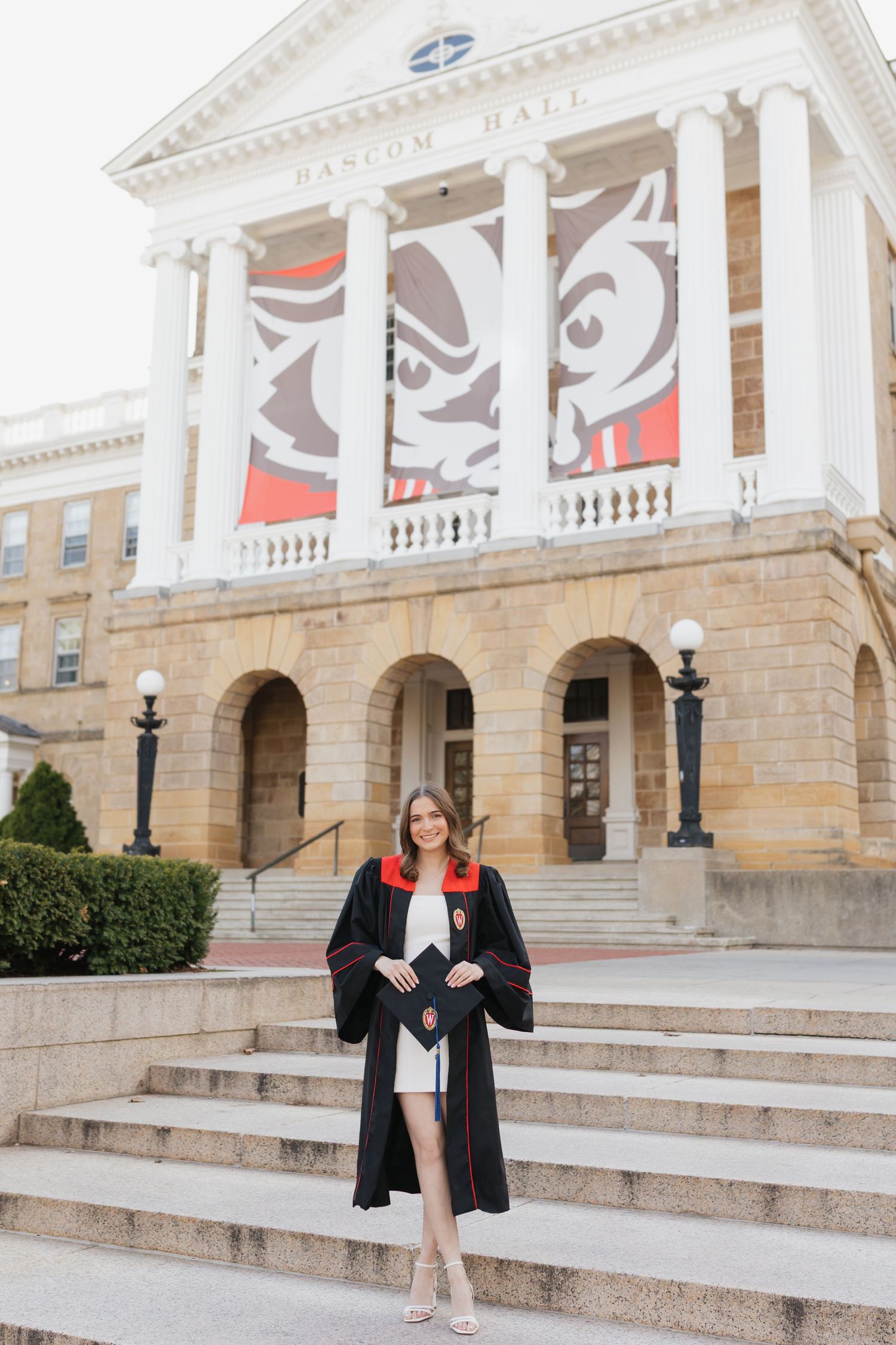 Graduate in black gown stands on steps of Bascom Hall with red and white banners.