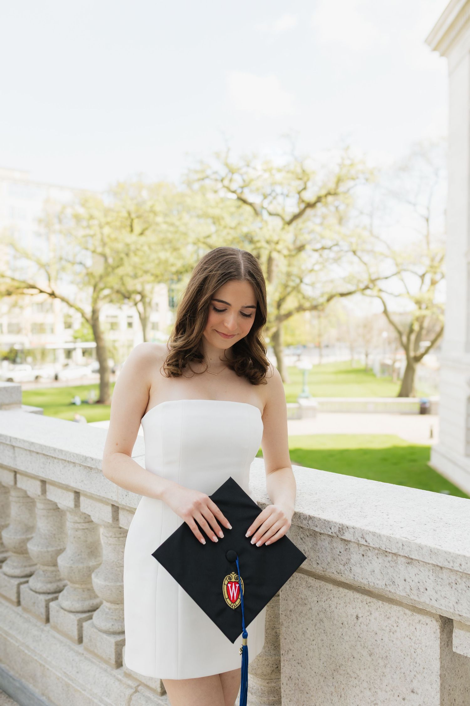 Graduate in white dress holds black diploma cover on stone balcony during spring.