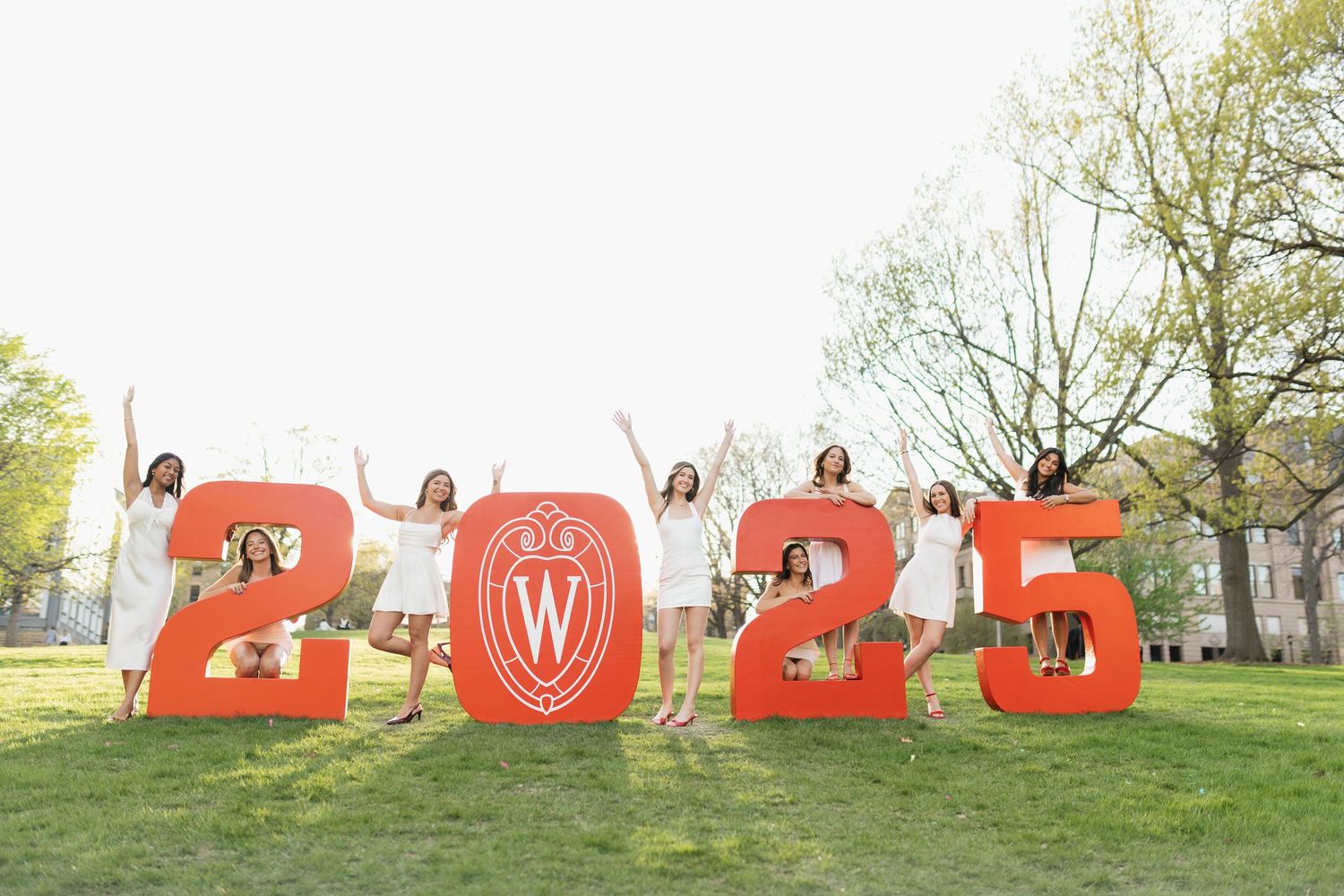 Graduates pose with red letter signs spelling out 2025 on a grassy hill.