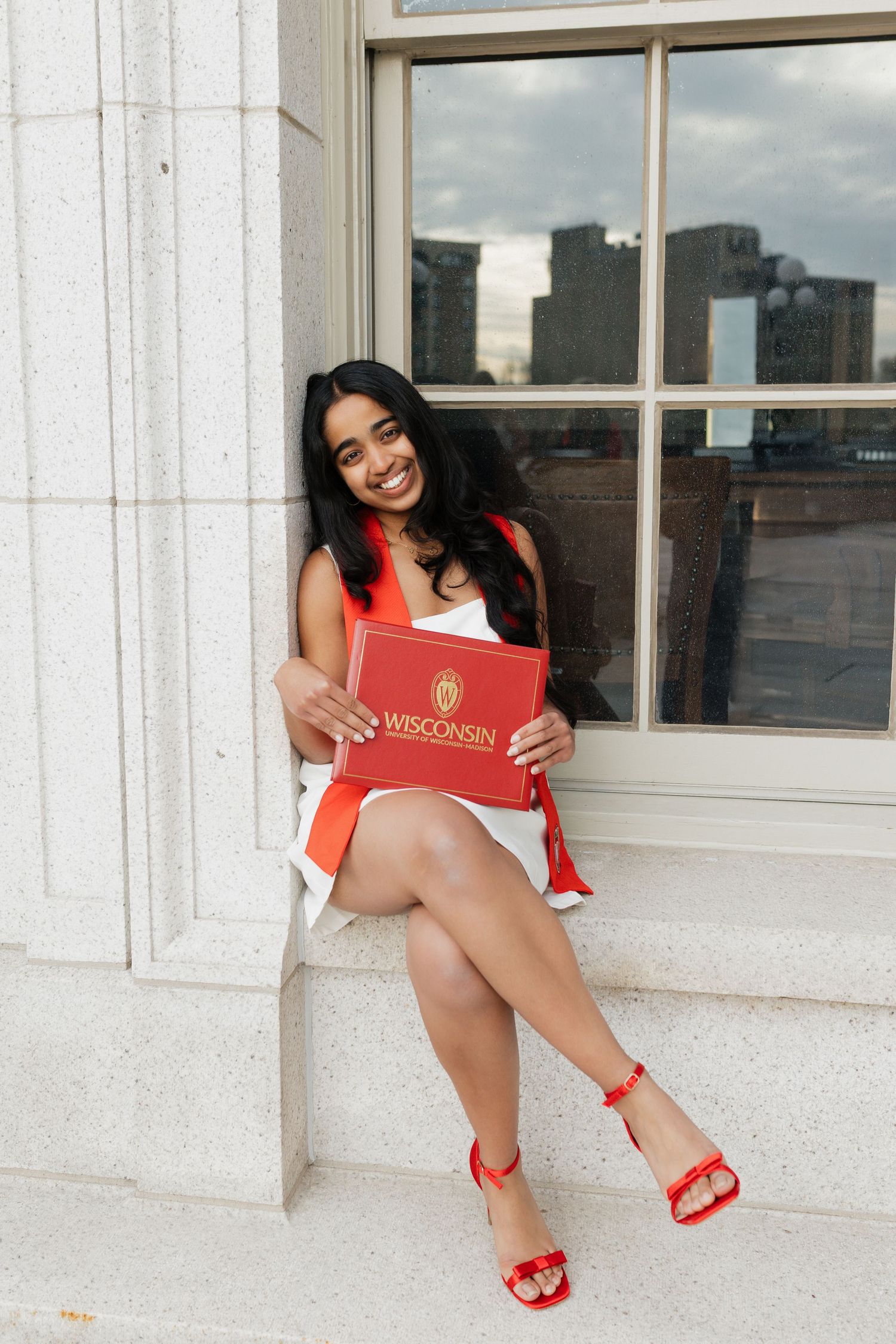 Graduate in white dress with red accessories poses by window ledge.