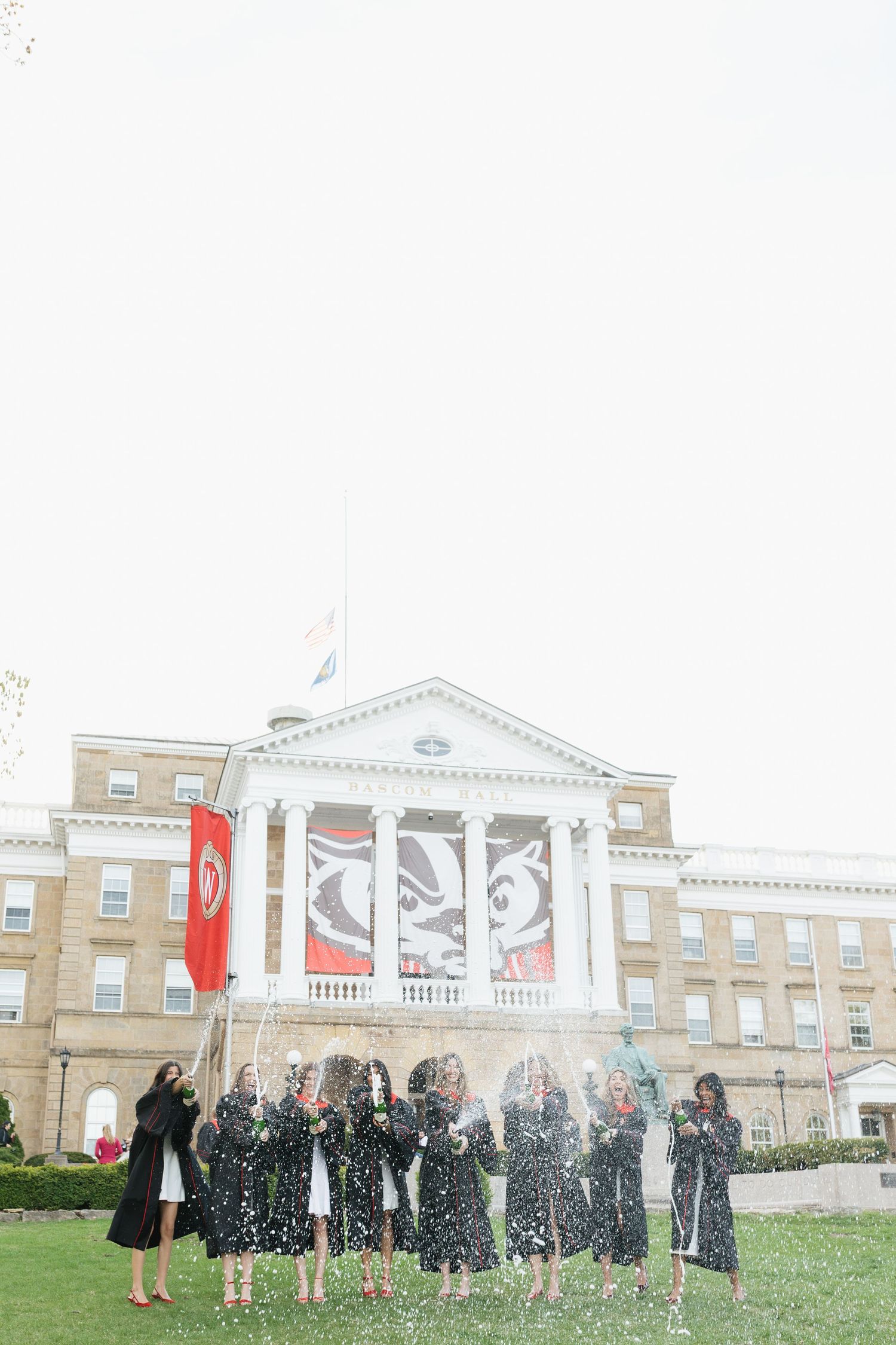 A group of graduates in black gowns celebrate by spraying champagne on a lawn in front of Bascom at UW Madison.