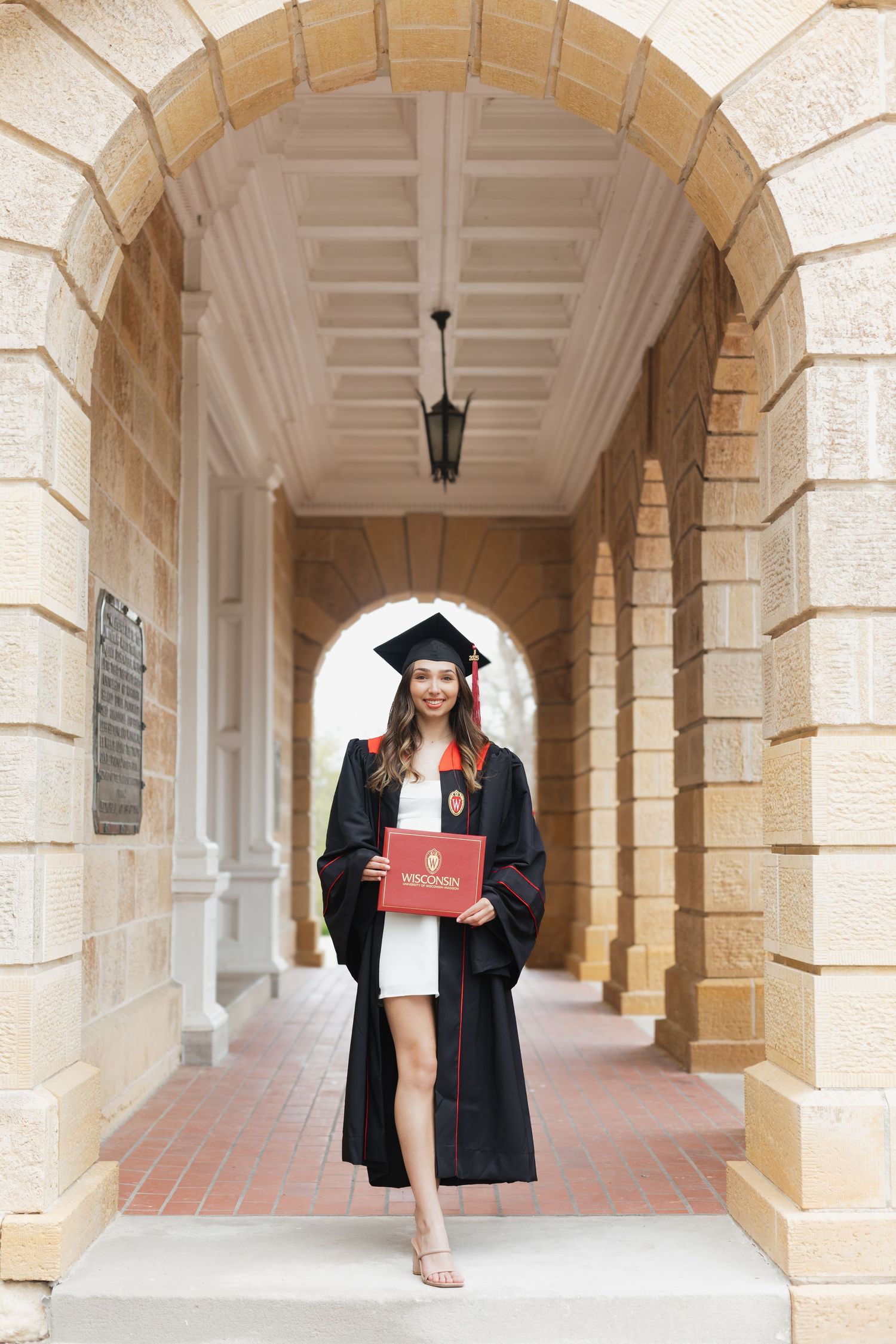 Graduate walks through arched hallway holding red diploma folder.