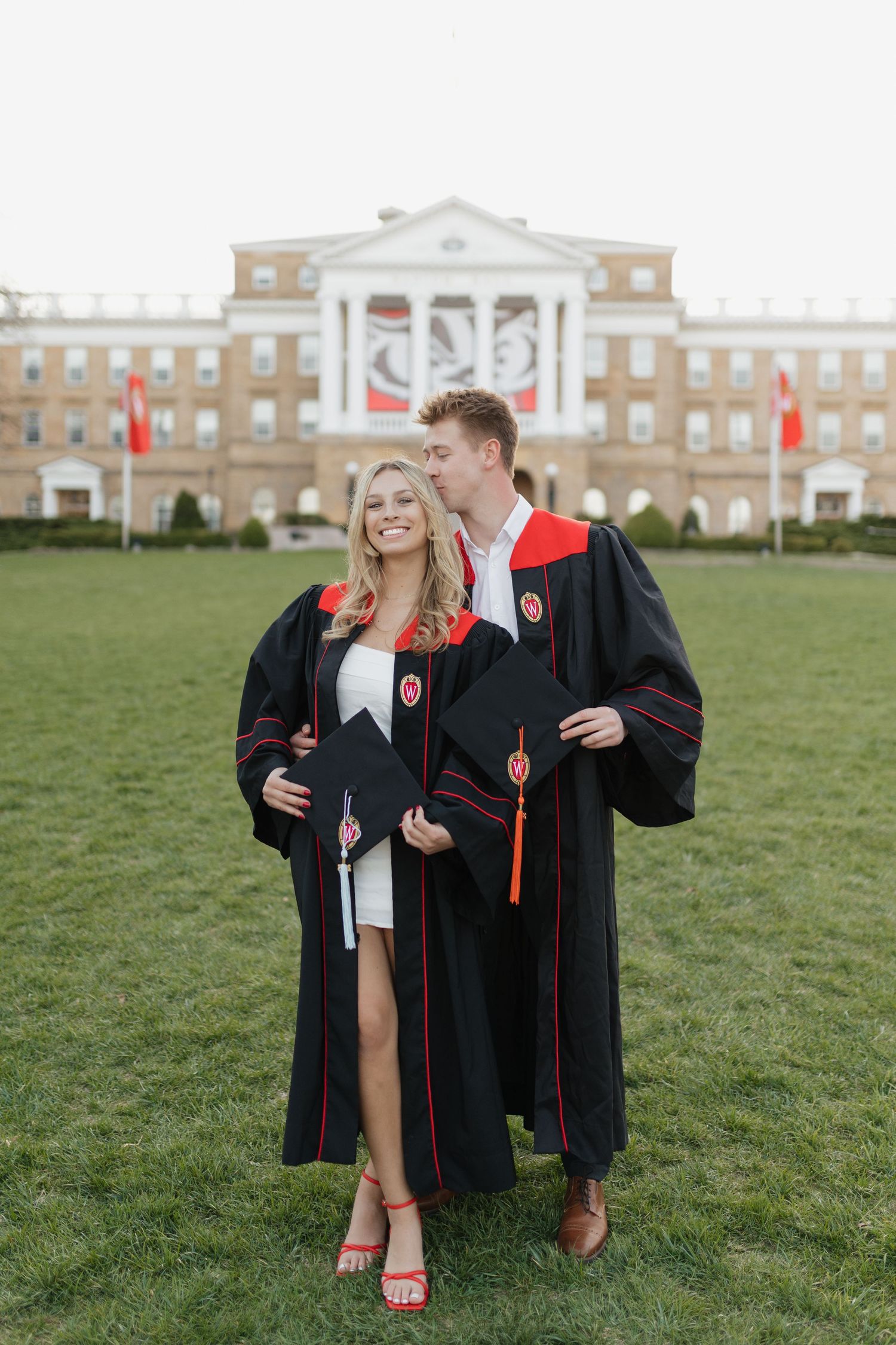 Graduates in black and red academic regalia pose on lawn in front of university building.