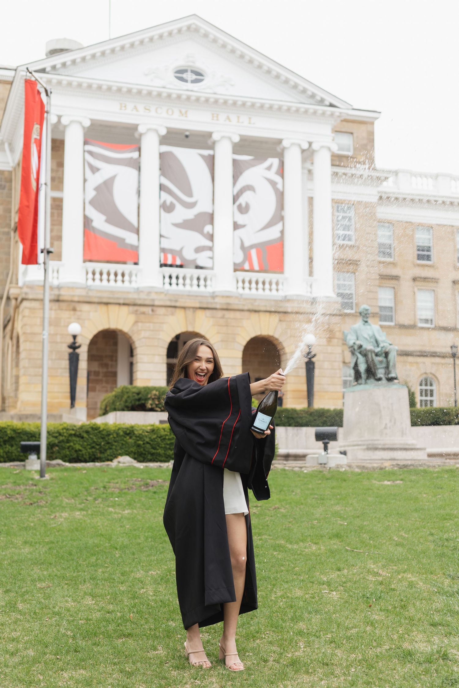 Graduate in academic regalia poses on lawn in front of grand university building.