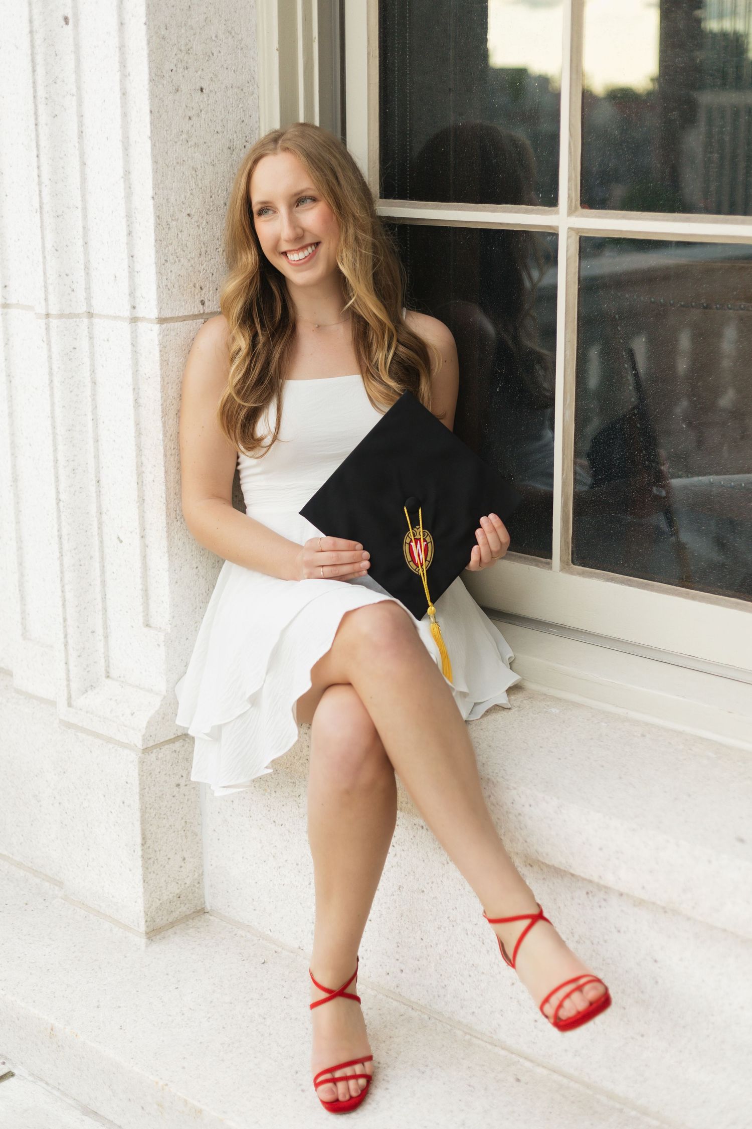 Graduate in white dress poses by column holding graduation cap.