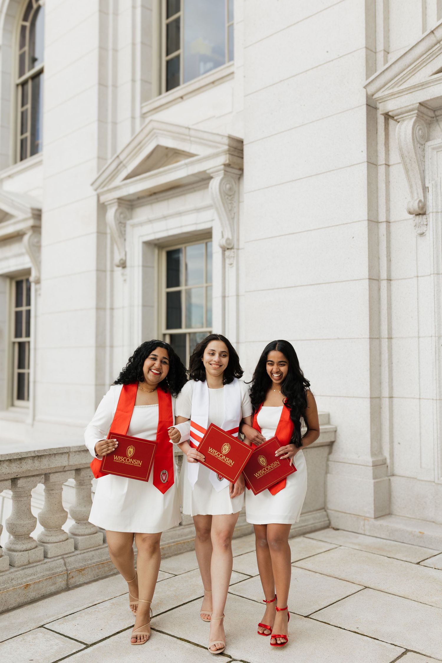 Three graduates in white dresses and red shawls pose together on a white stone balcony.