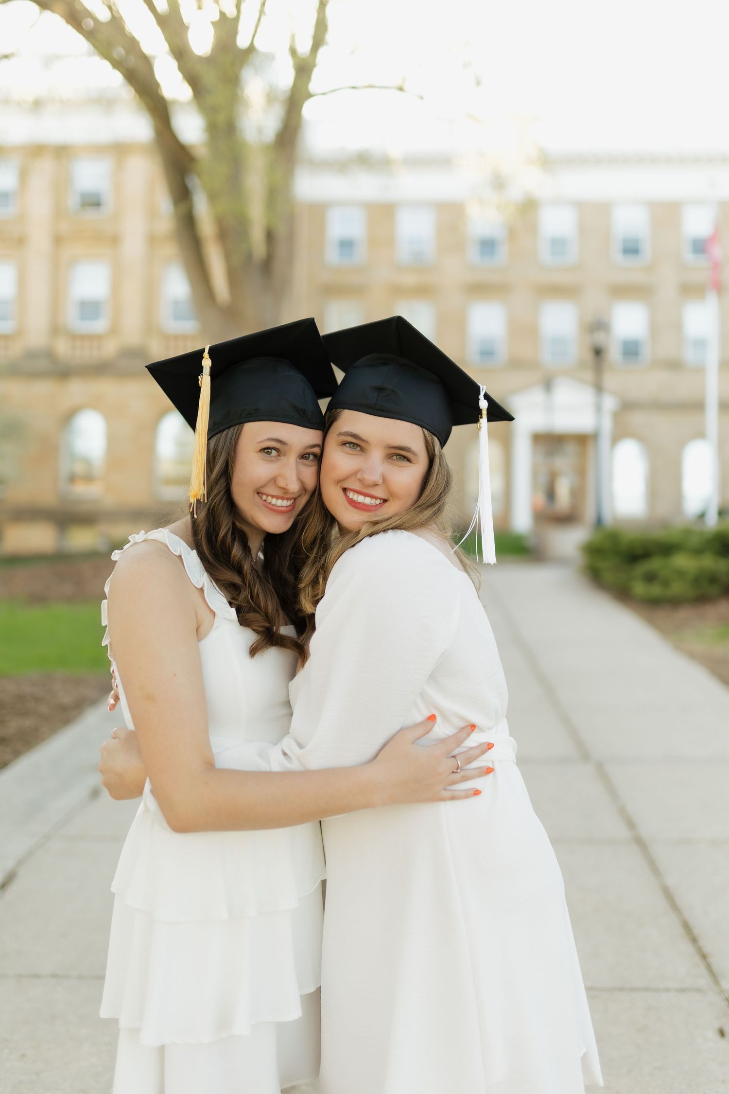 Two graduates in white dresses and caps embrace while celebrating on campus grounds.