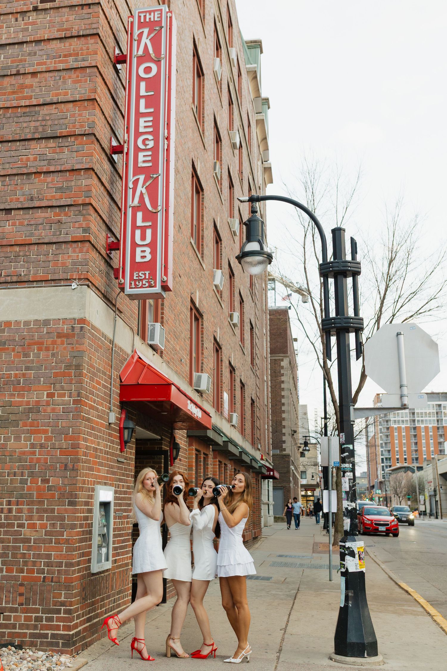 UW Madison graduates in white dresses and red heels poses along brick wall of urban building with vintage-style lamp posts.