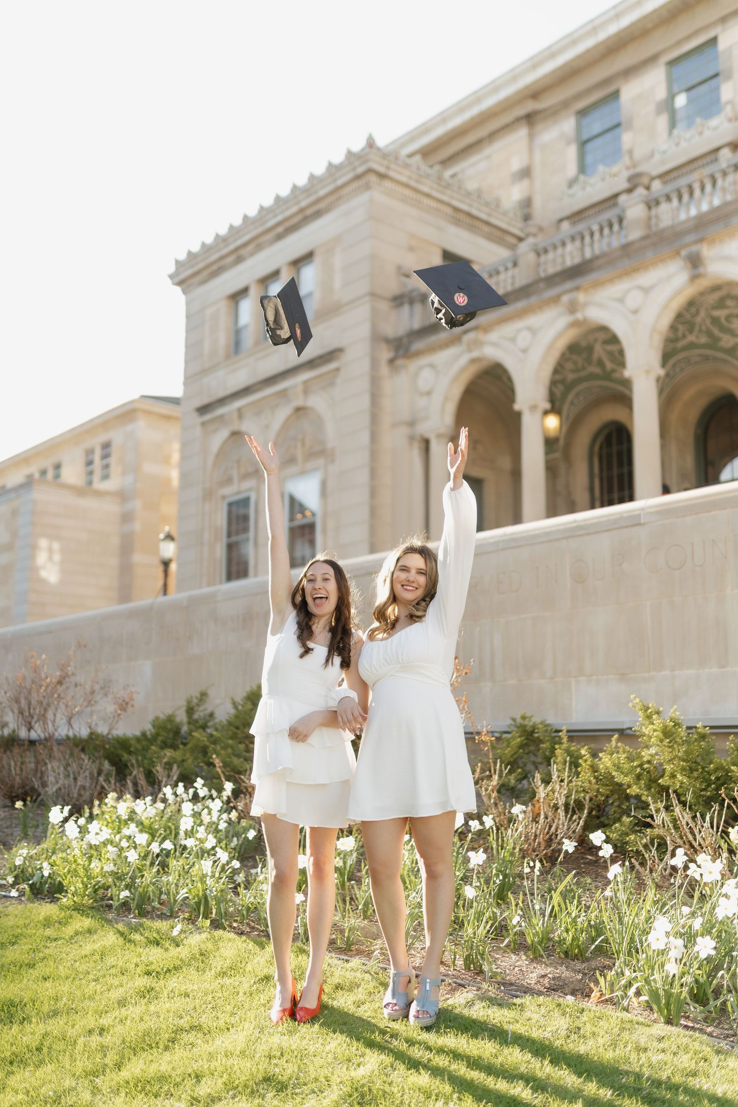 Graduates in white dresses celebrate among spring flowers on university grounds.