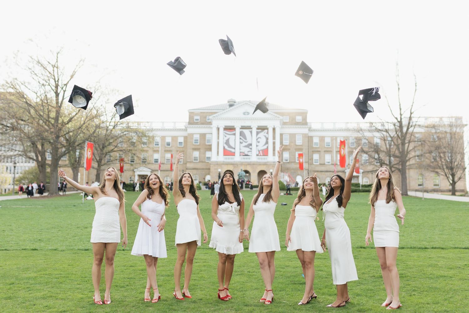 Graduates in white dresses line up on a grassy lawn for a celebratory photo.
