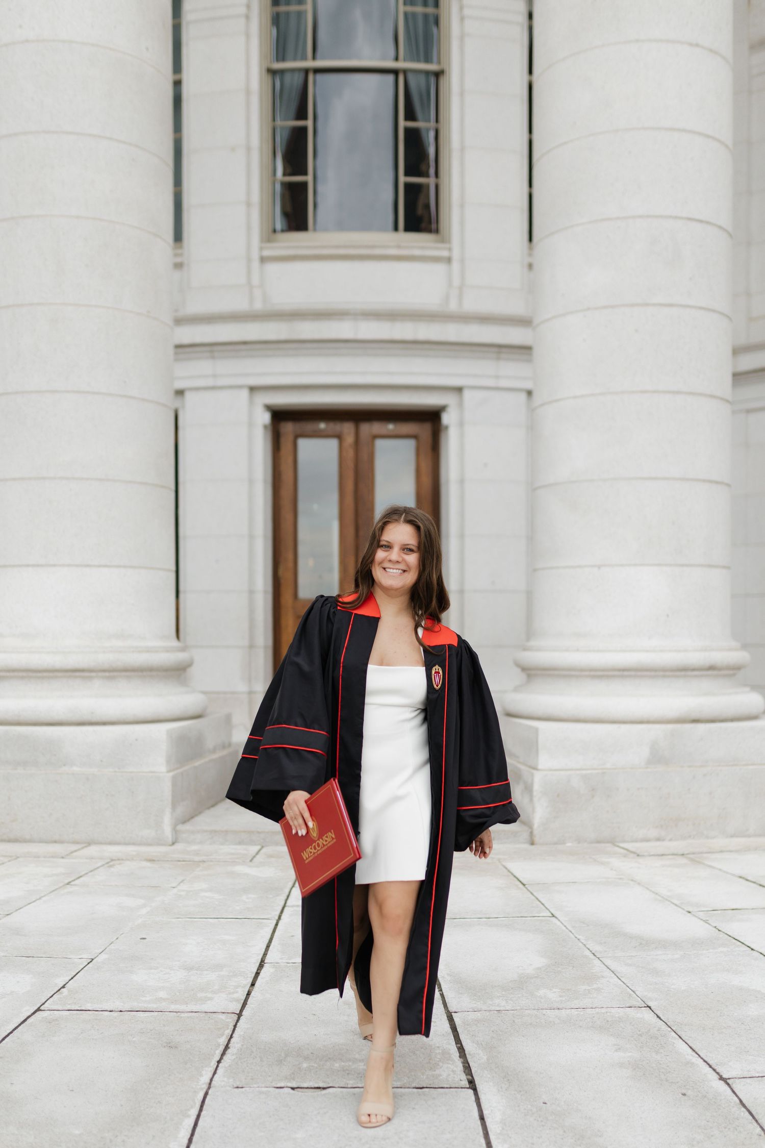Graduate in black and red graduation gown poses against white stone columns.