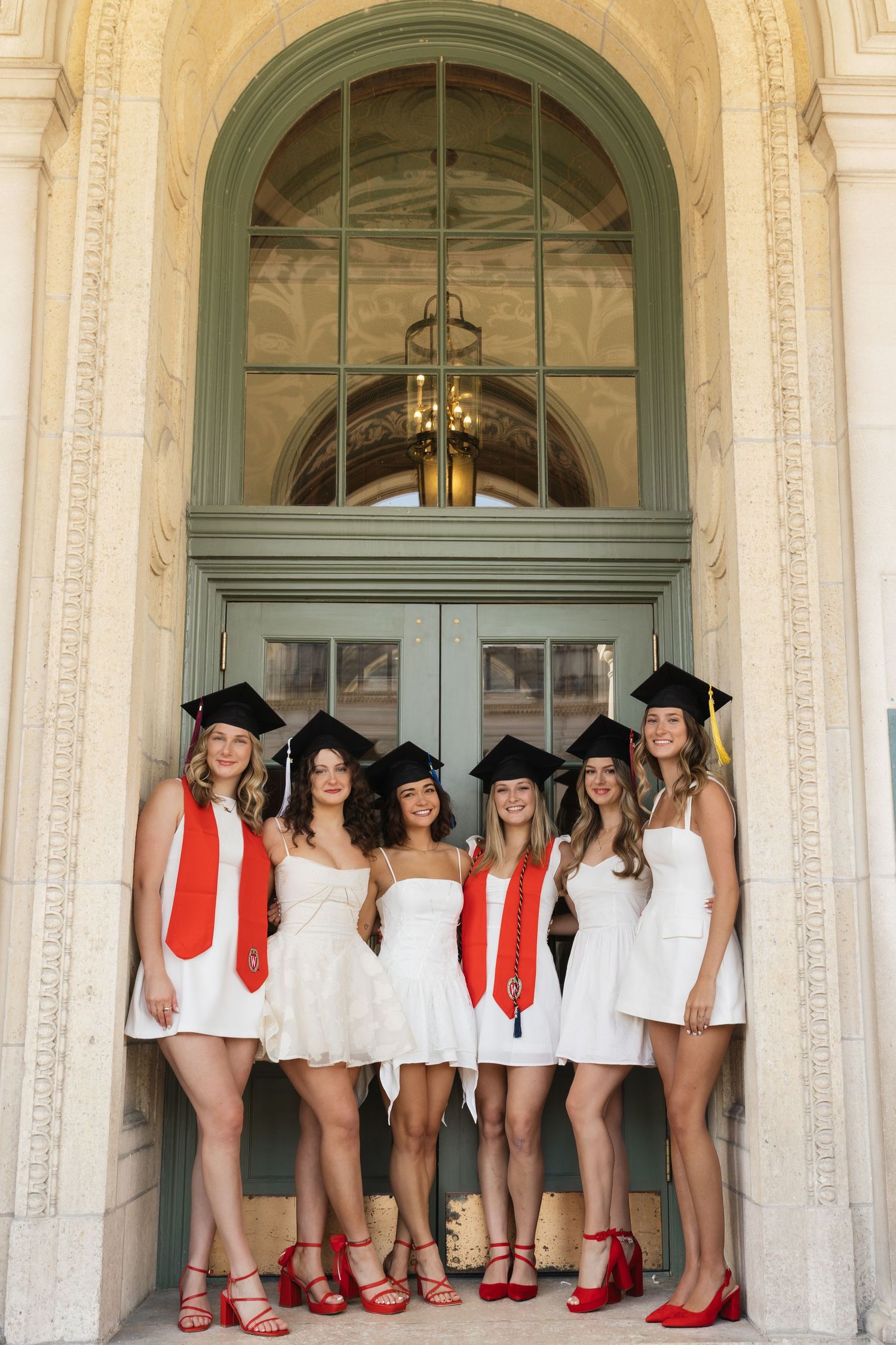 A line of graduates in white dresses and red shoes pose together under ornate architecture.