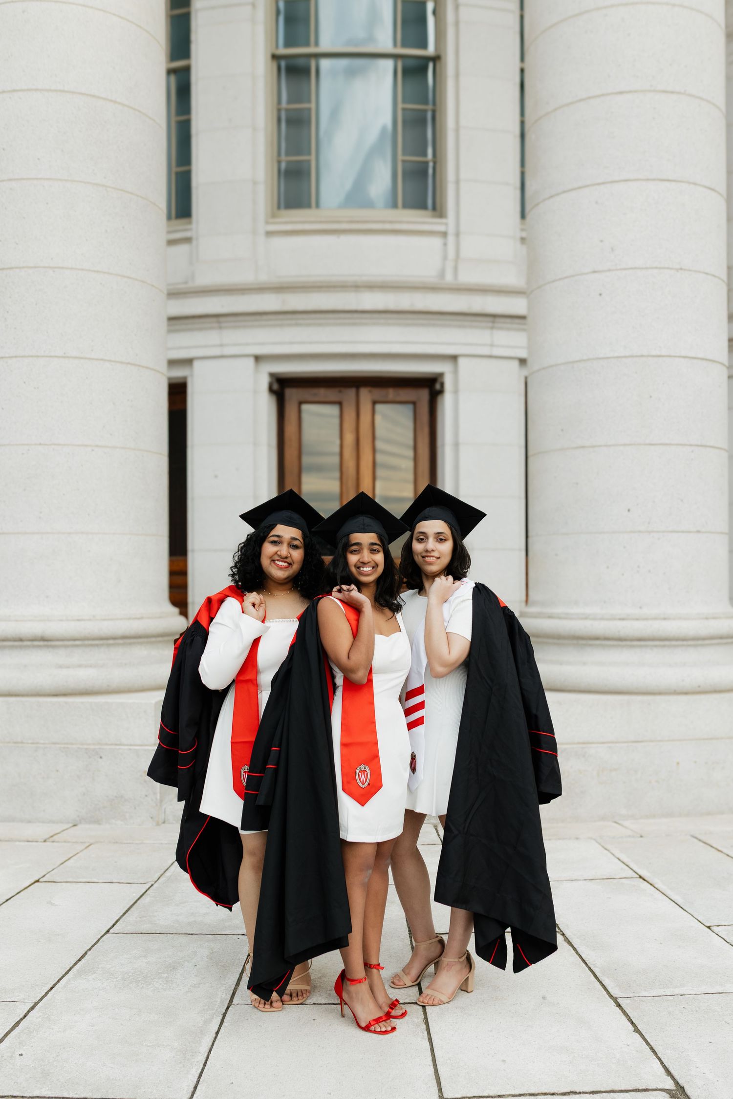 Graduates wearing black caps and gowns pose on a classic white stone building exterior.