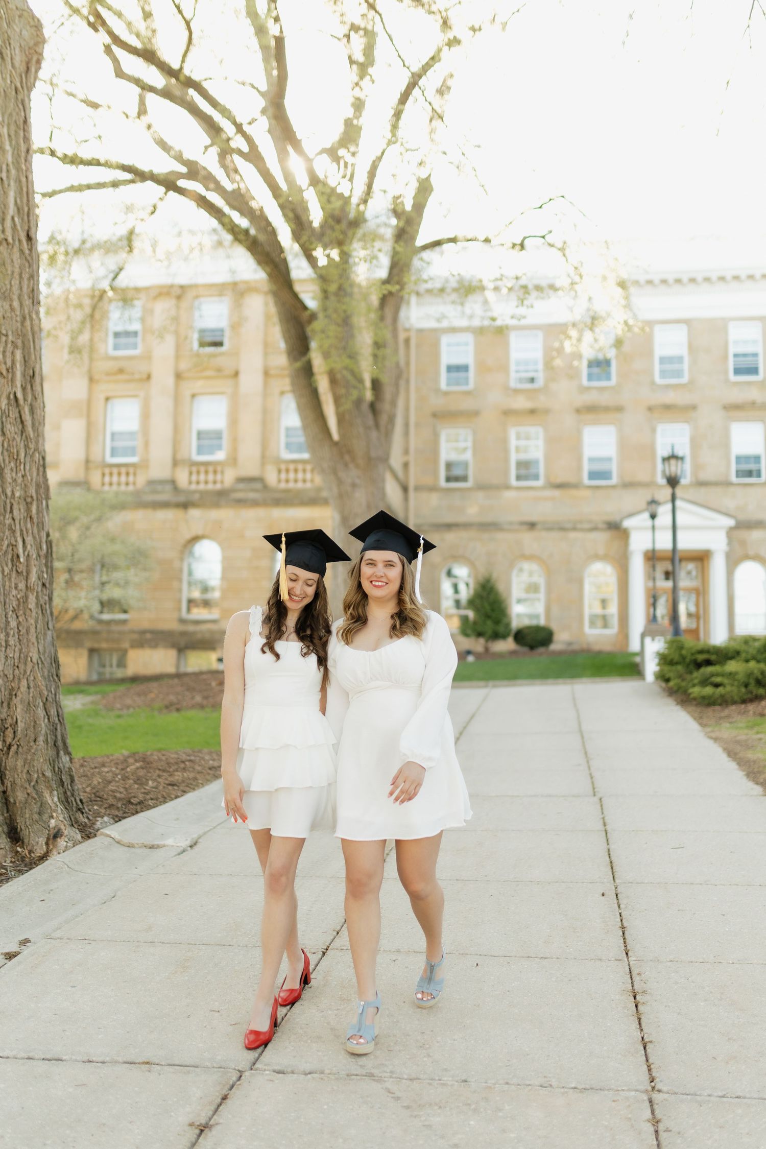 Graduates in matching white dresses walk along a campus sidewalk with historic buildings in background.