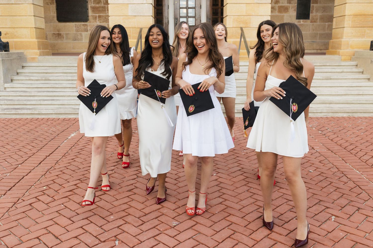 Graduates in white dresses and black graduation gowns walk along a brick pathway.