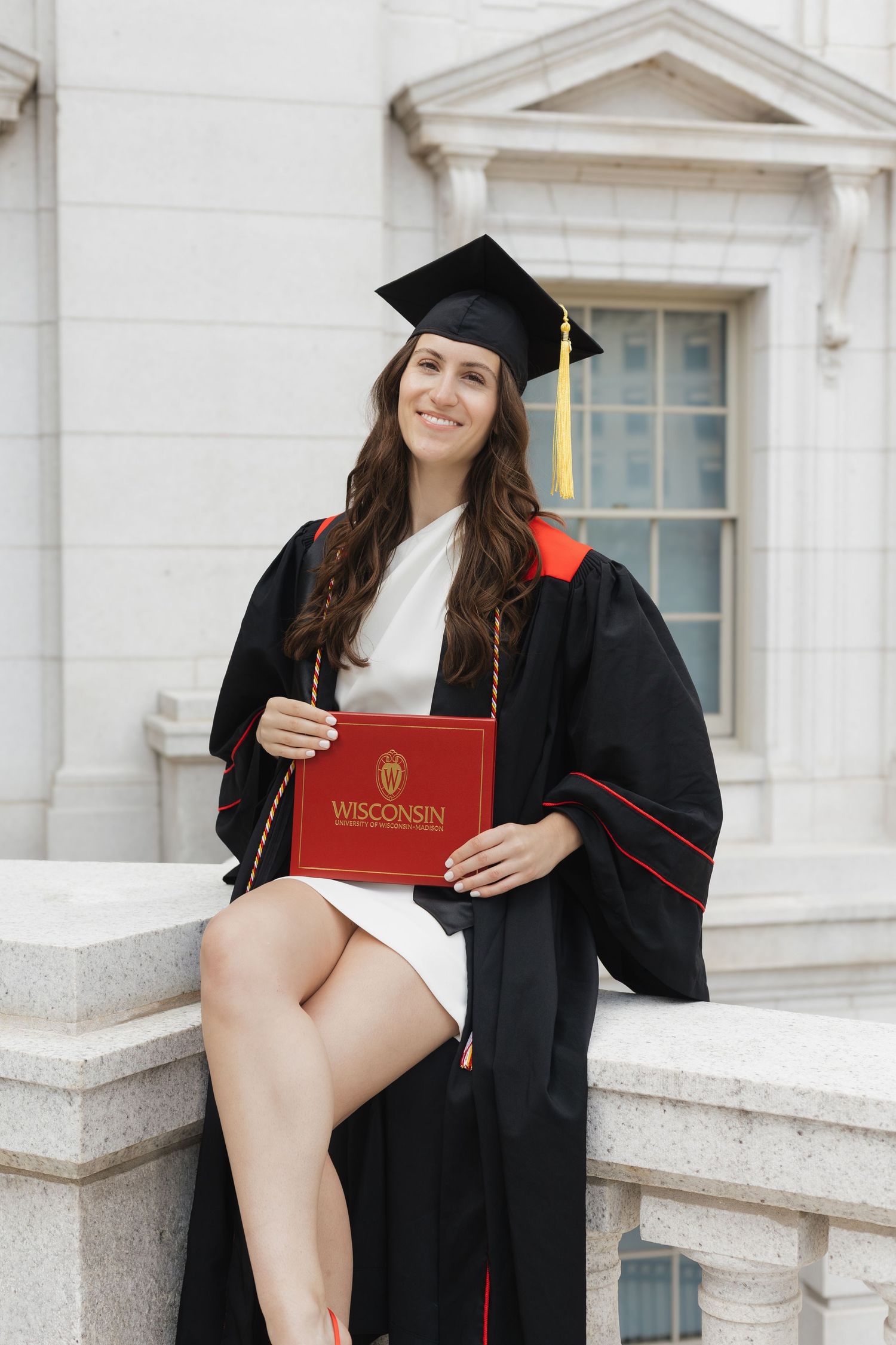 Graduate displays University of Wisconsin diploma while sitting on stone steps.