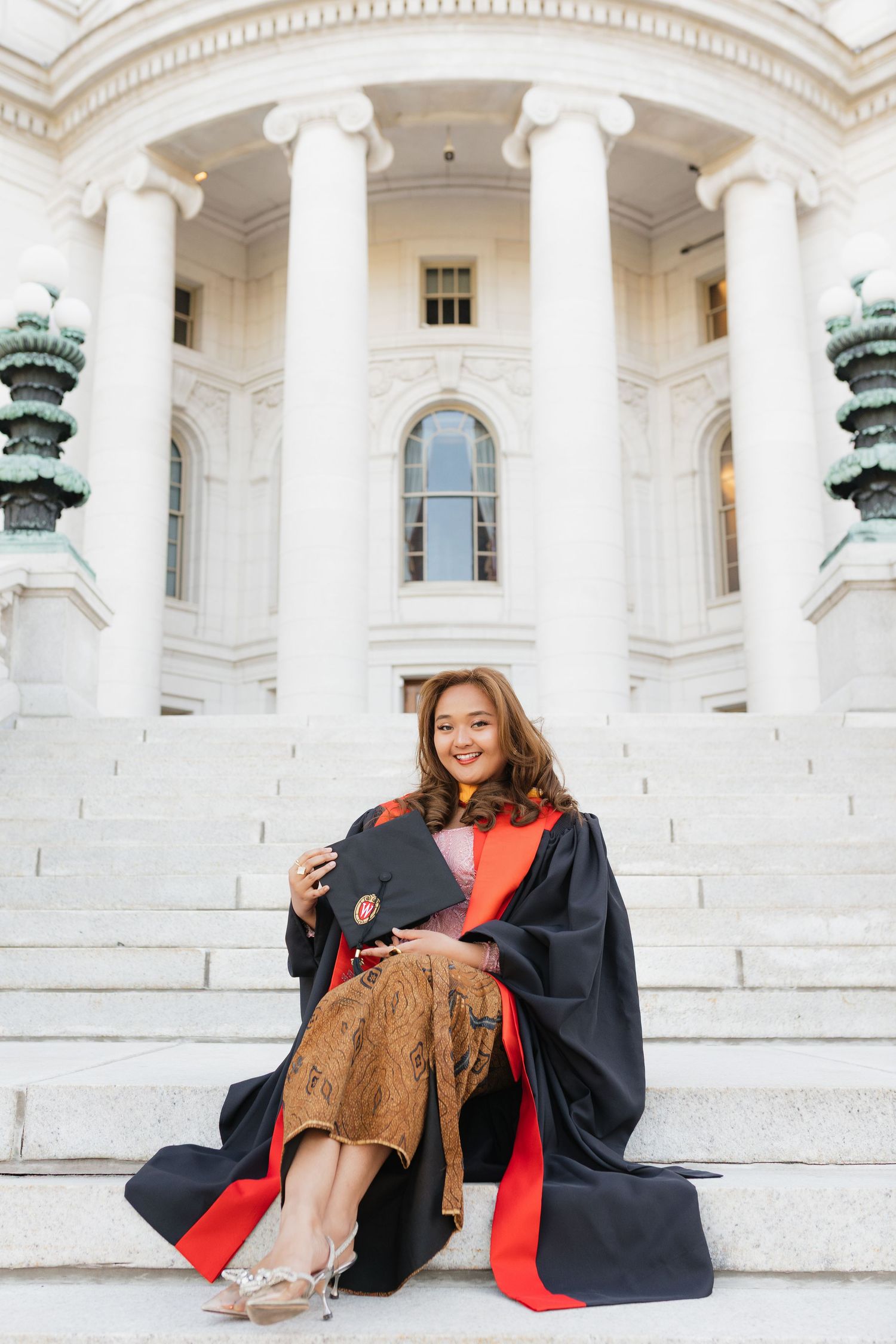 Graduate in ceremonial robes sitting on grand white steps with classical architecture.