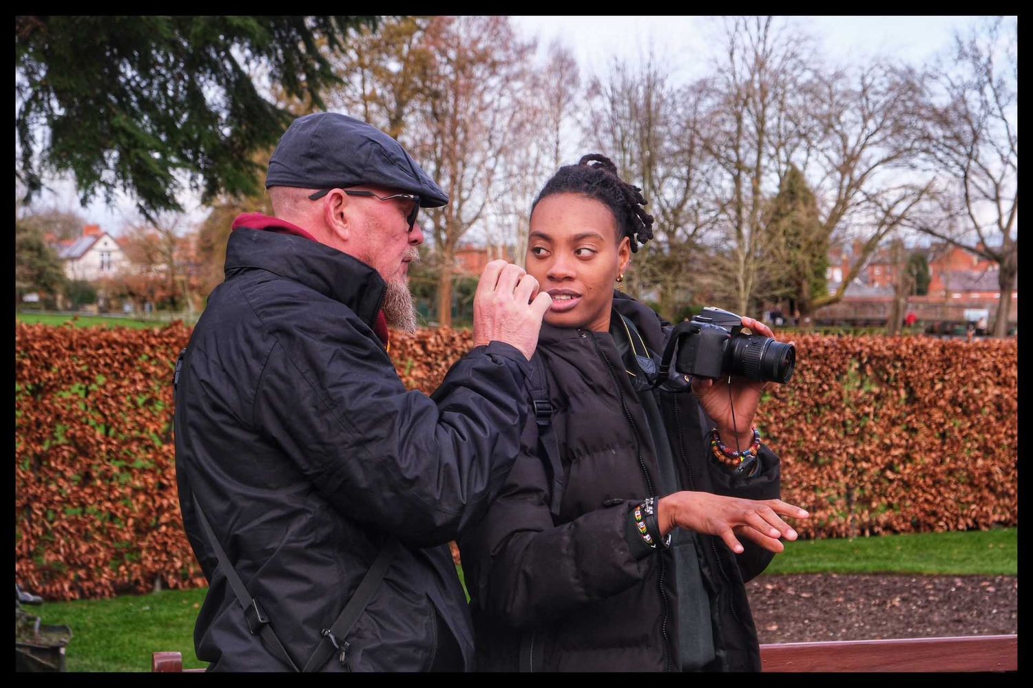 Rob Kirman and a student stand outdoors examining a camera near an autumn hedge in a park.
