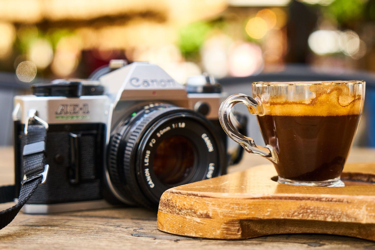 A Canon film camera sits beside a glass cup of black coffee on a wooden board.