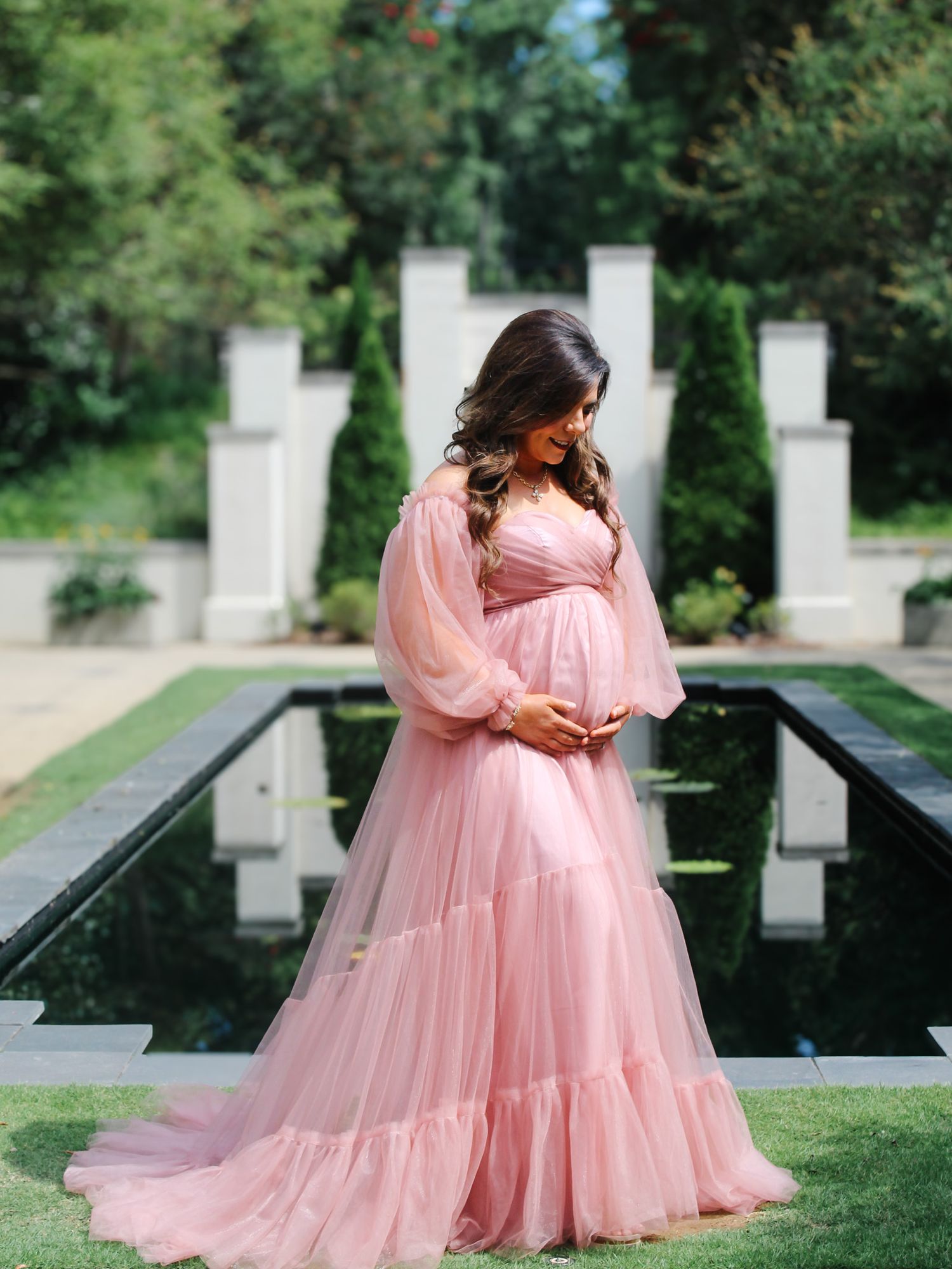 A flowing pink maternity gown photographed by a reflecting pool with white pillars in the background.
