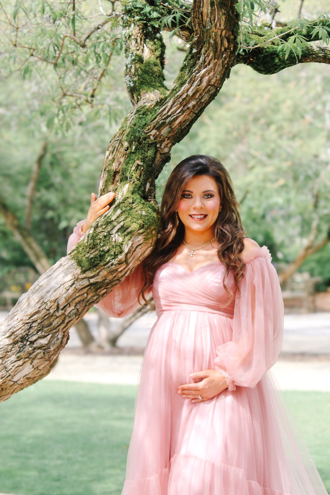 A maternity photoshoot in a garden setting with a flowing pink tulle gown against a rustic tree backdrop.