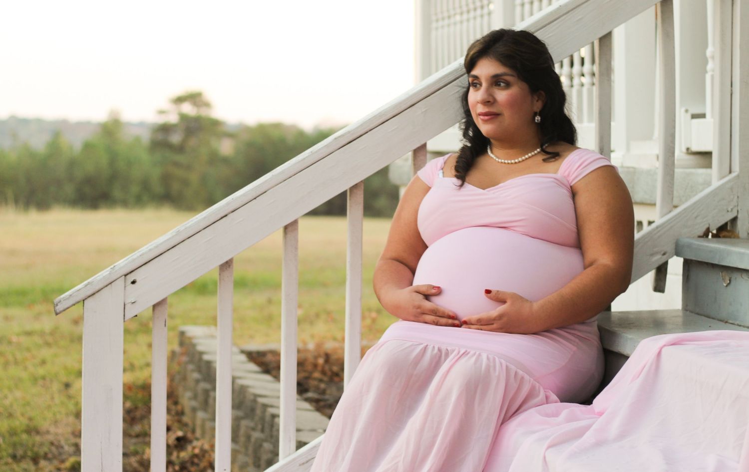 A person in a pink maternity gown stands on white porch steps during an outdoor photo session.