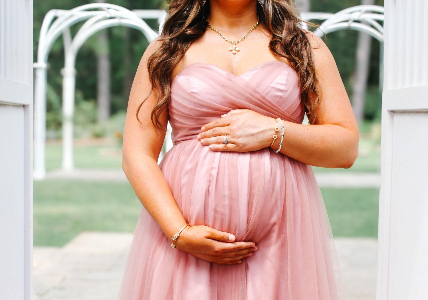 Woman in flowing pink tulle gown poses gracefully in outdoor garden setting.