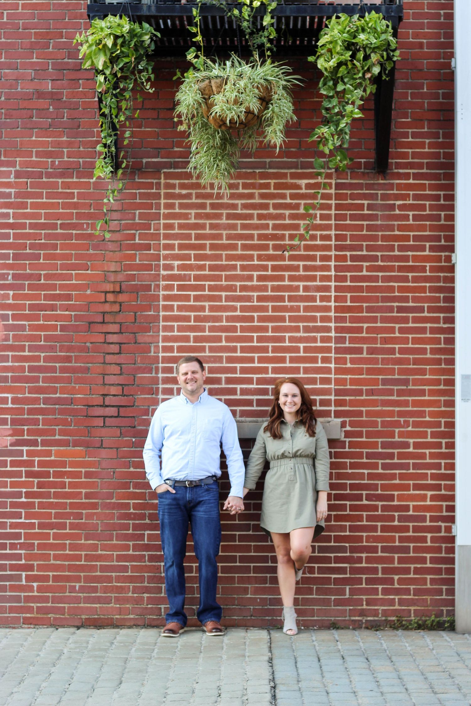 Couple leans against red brick wall with hanging plants.