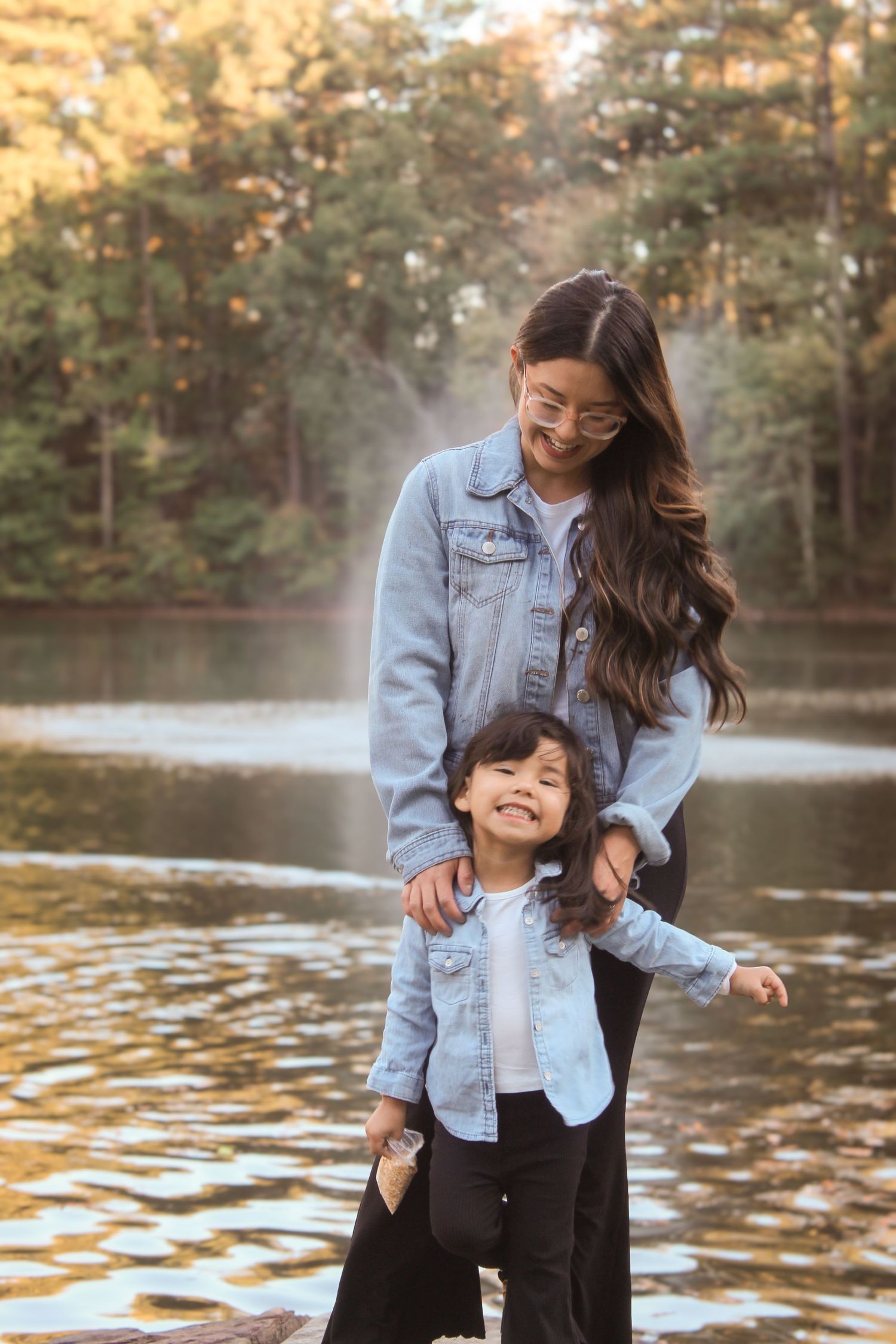 Mother and child sharing tender moments by a lake in denim jackets during sunset.
