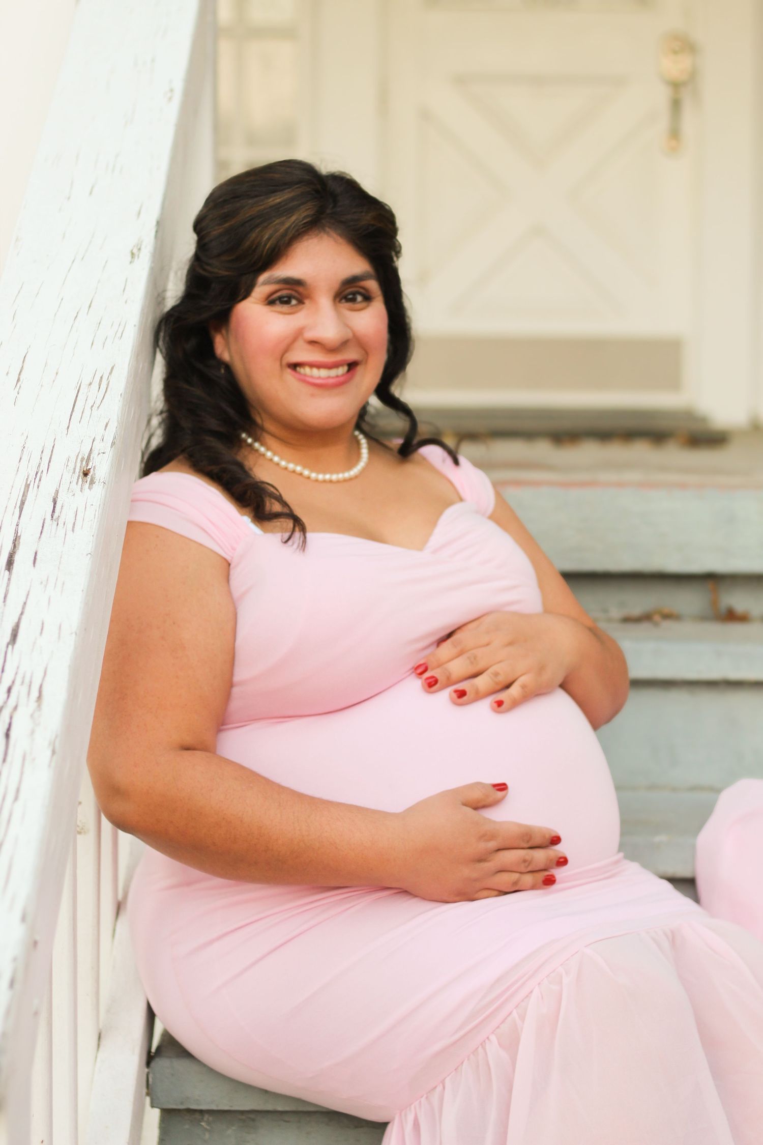 A person in a pink dress sits on white stairs during a maternity photoshoot.