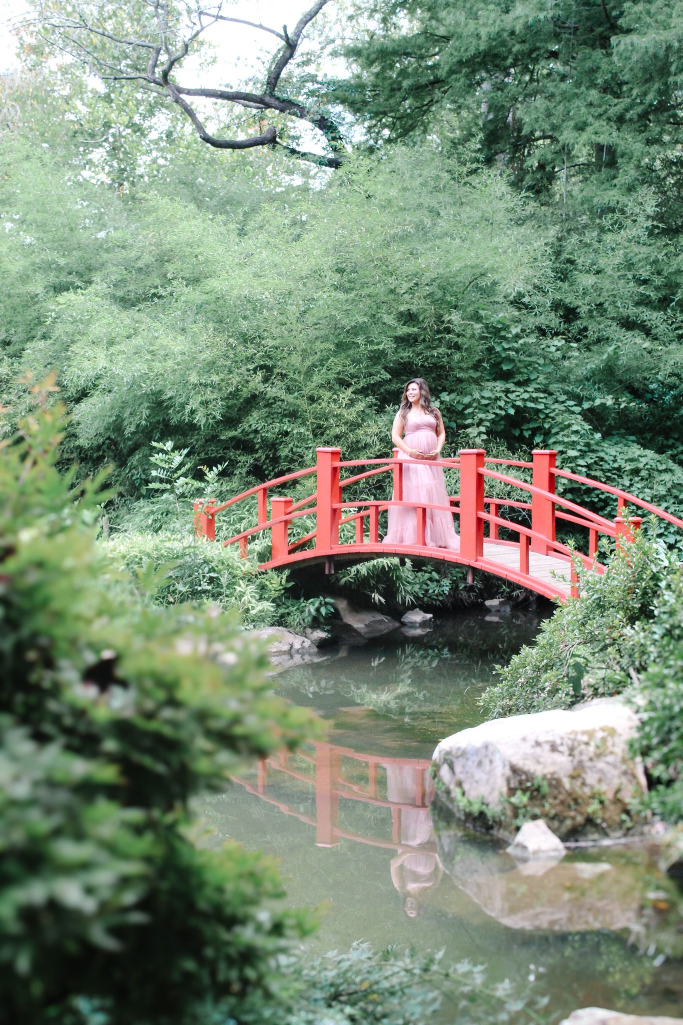 Red Japanese bridge spans across peaceful pond in lush garden setting.