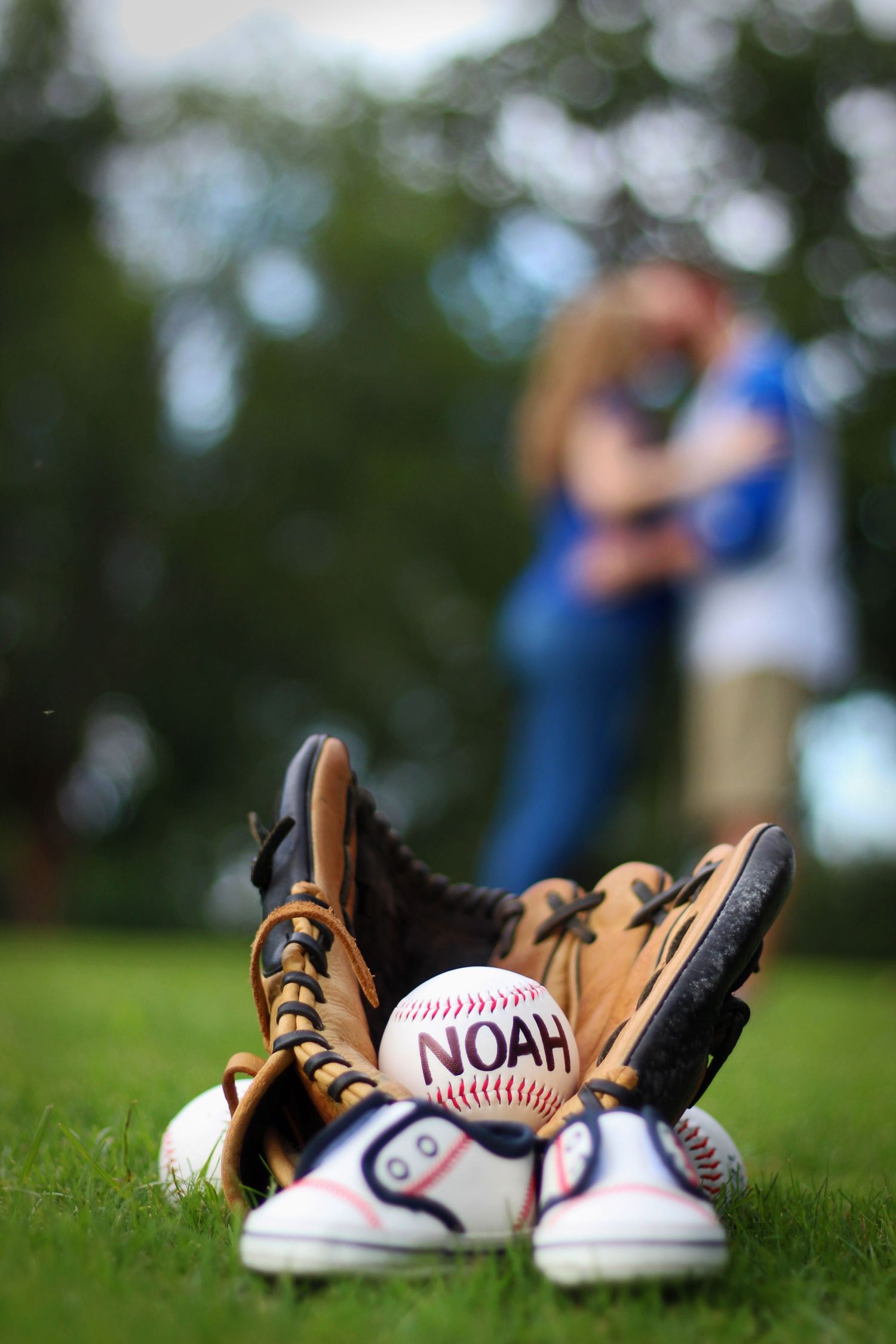 Baseball glove and balls with name NOAH arranged on grass with blurred figures in background.