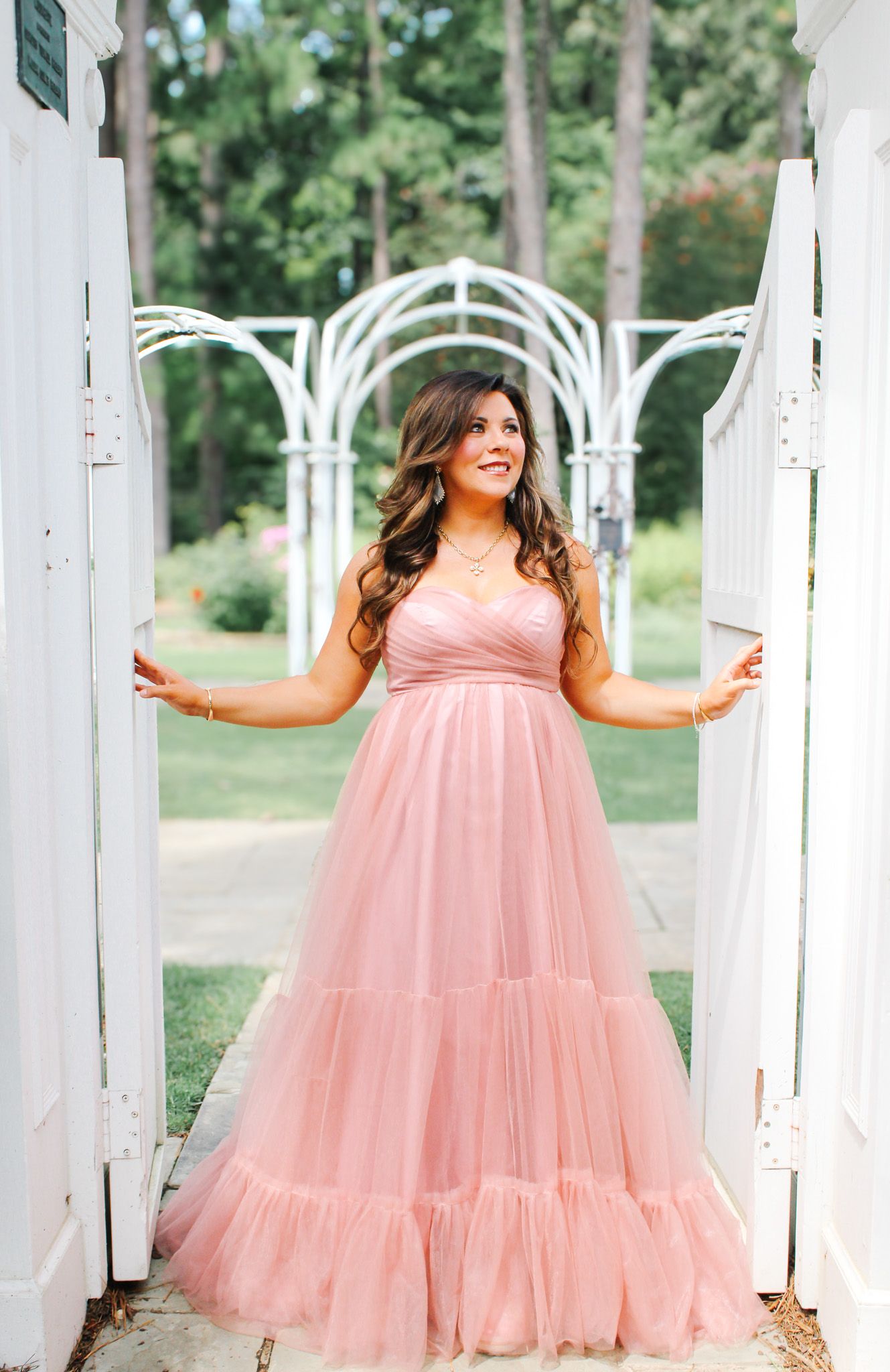 A joyful maternity portrait session featuring a flowing pink dress against white garden pillars.