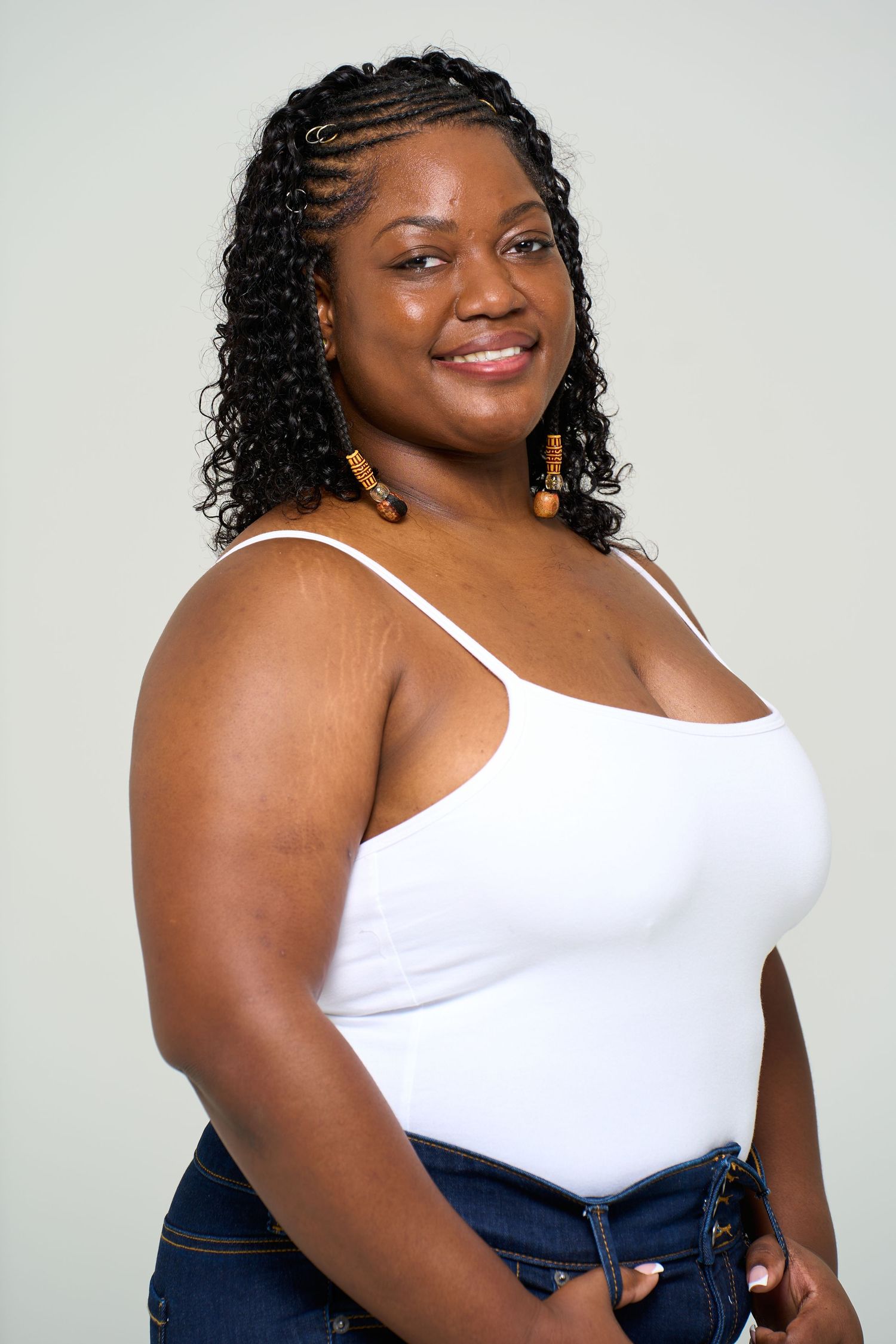 Confident woman in white tank top and jeans poses against light background in studio photoshoot.
