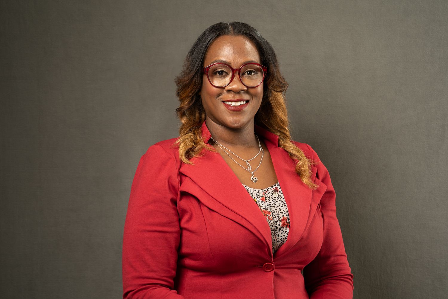 Professional headshot of someone in a red blazer and glasses against a gray backdrop.