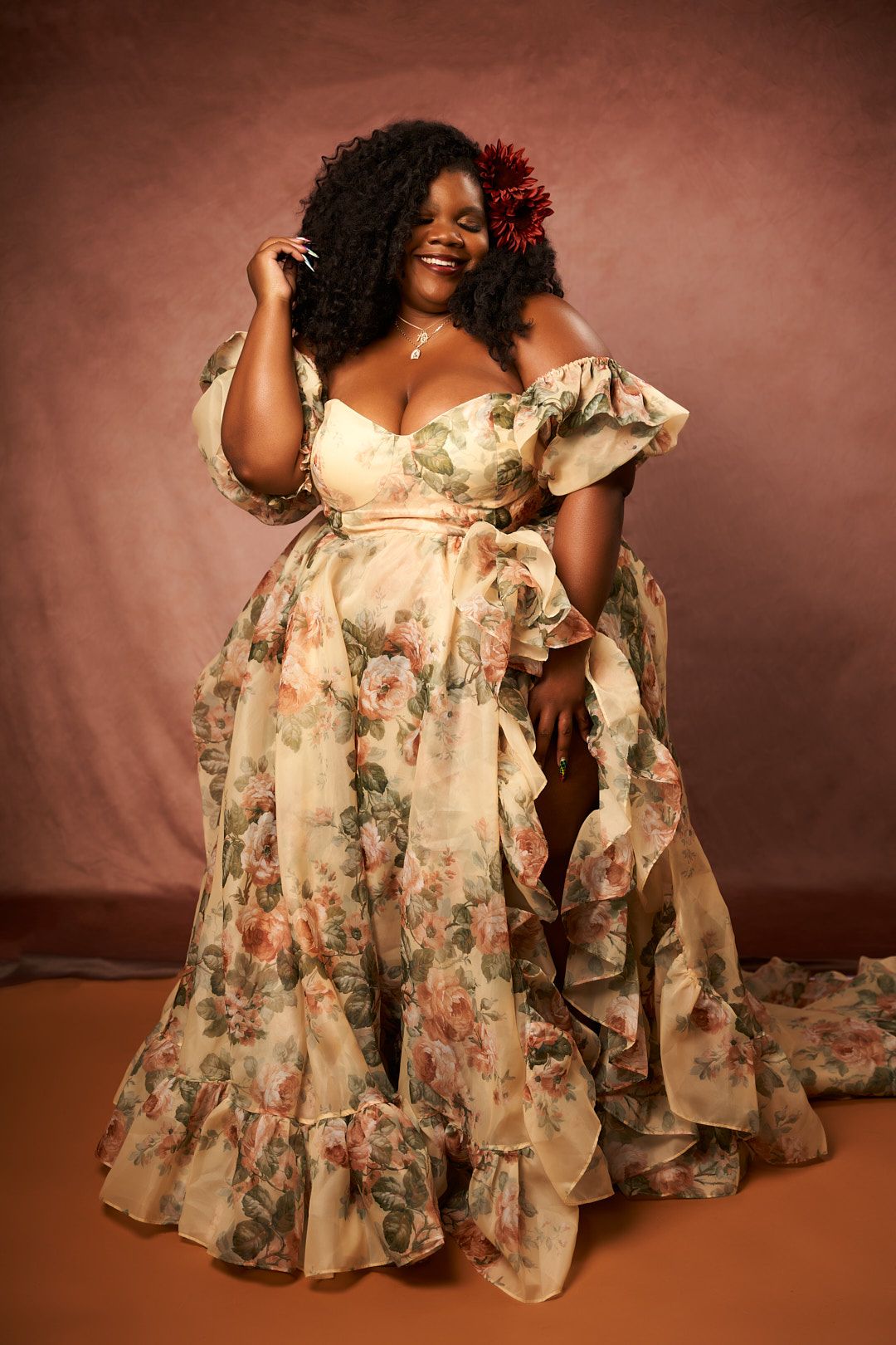 A floral print chiffon dress with ruffled details flows elegantly against a warm brown studio backdrop.