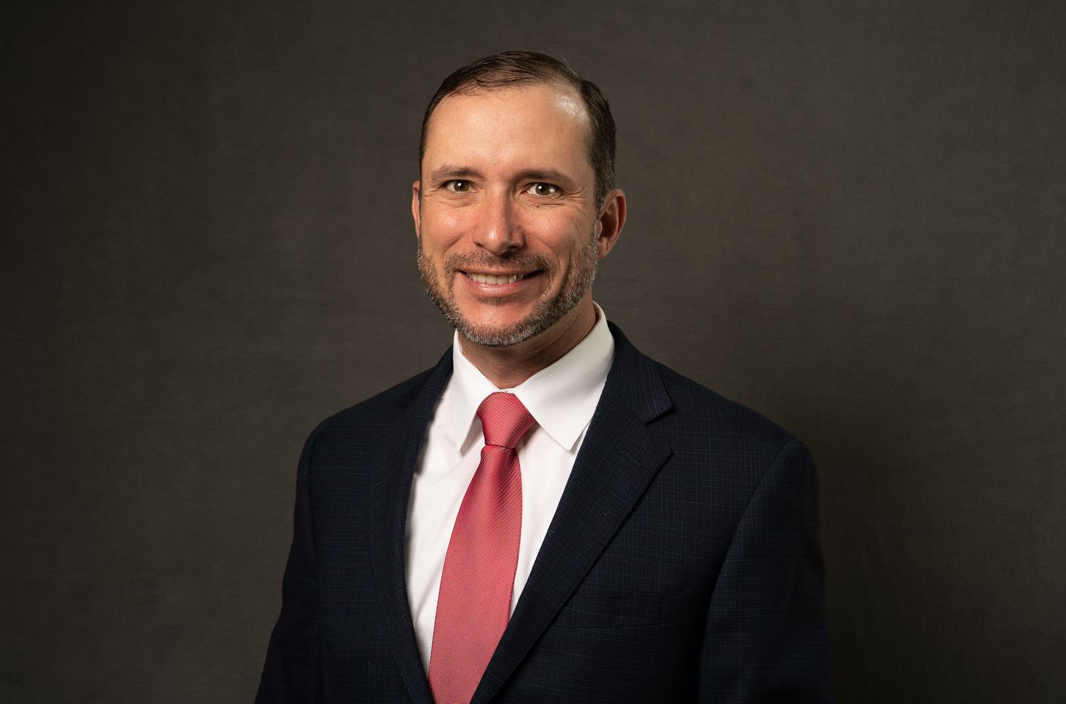 Professional headshot portrait of someone in a dark suit and red tie against a gray studio background.