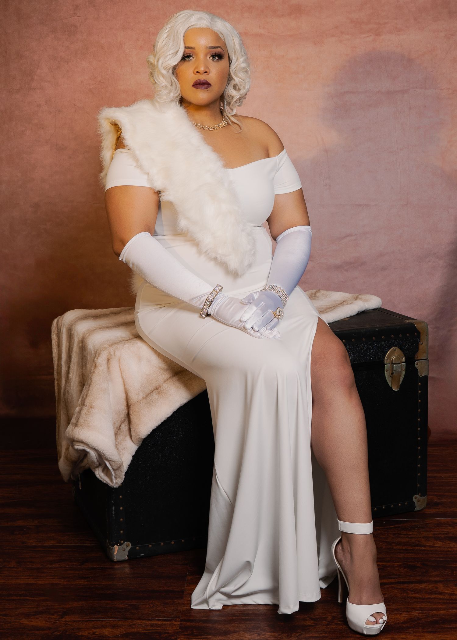 A person in a glamorous white dress and gloves poses elegantly on a dark chair against a textured mauve backdrop.