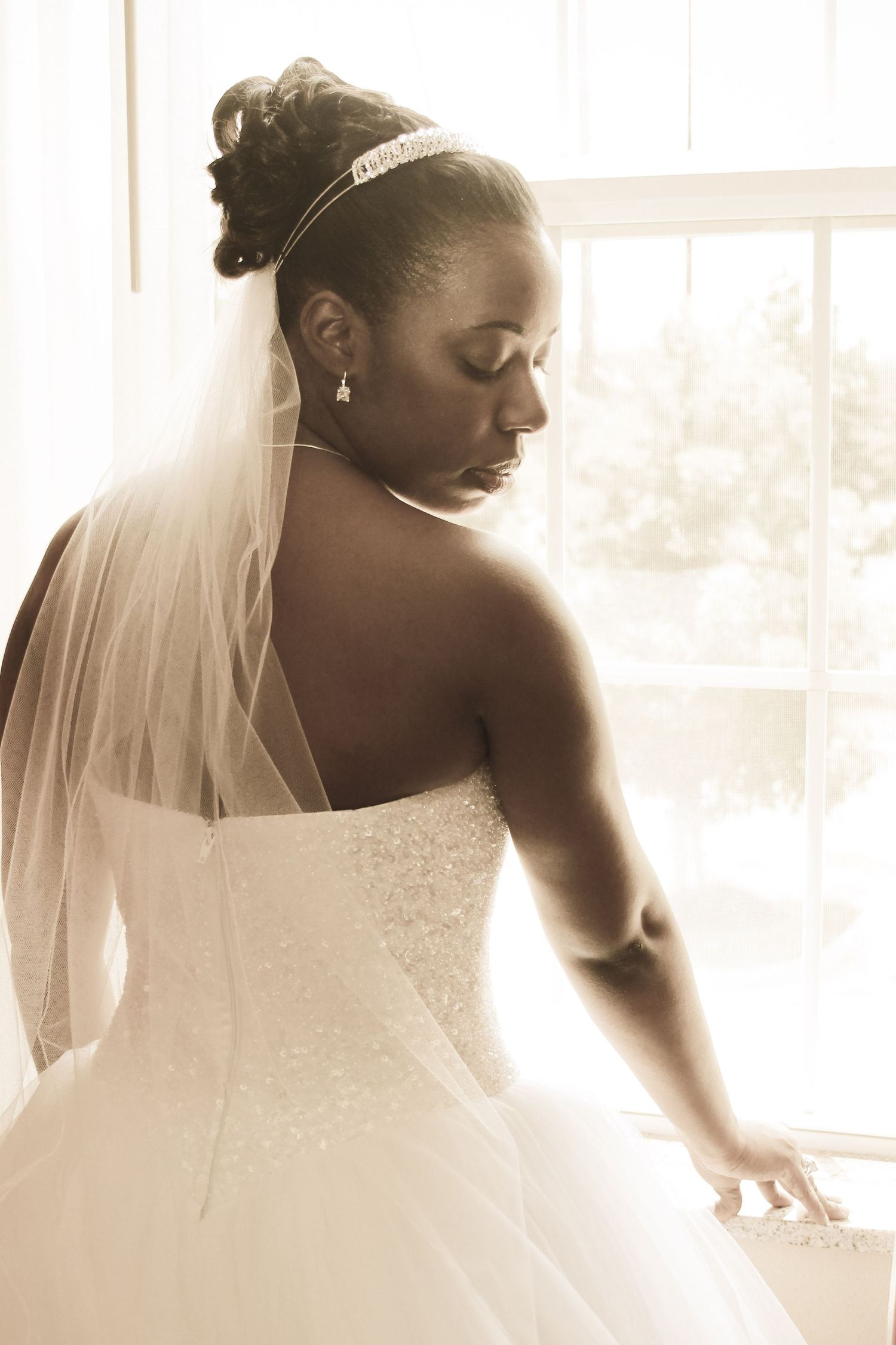 A bride in a white wedding dress and veil poses by a sunlit window in a romantic sepia-toned photo series.