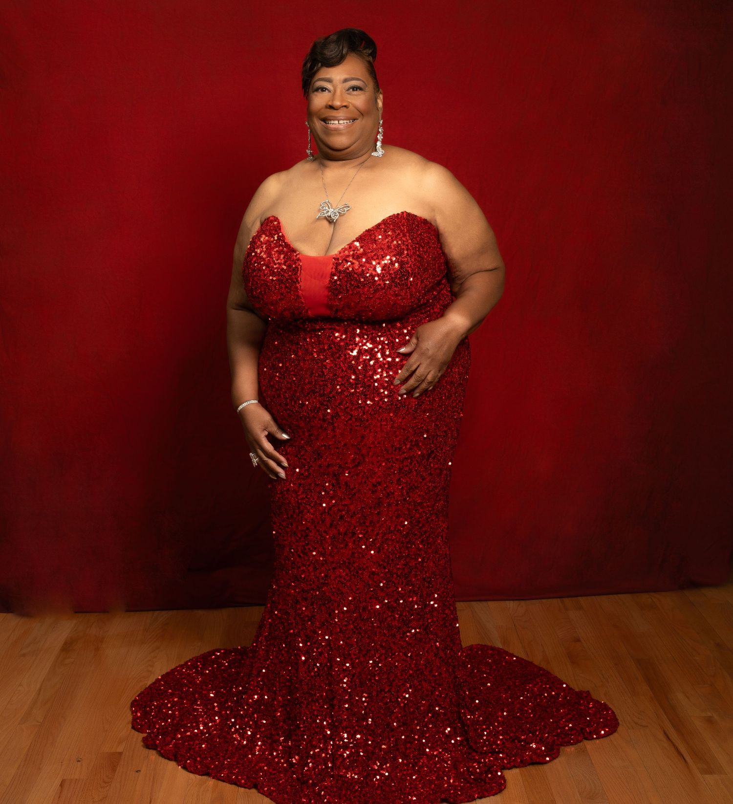 A person in a sparkling red evening gown poses against a dark burgundy background in a glamorous photoshoot.