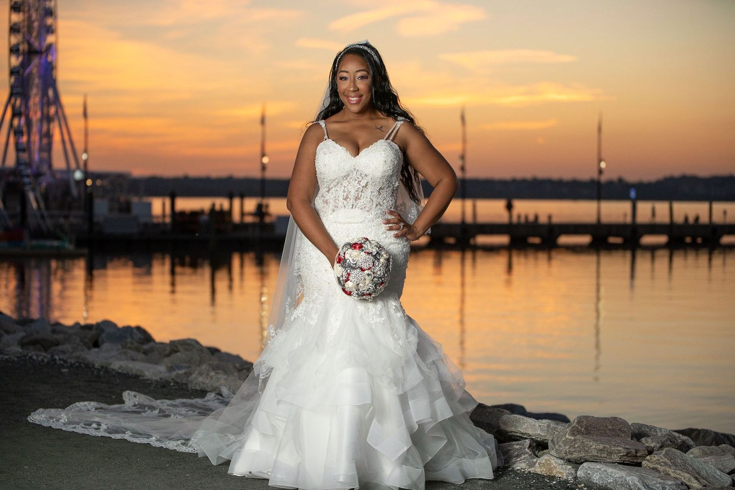 A bride in a white wedding gown poses against a stunning orange sunset over a marina with boats and docks in the background.