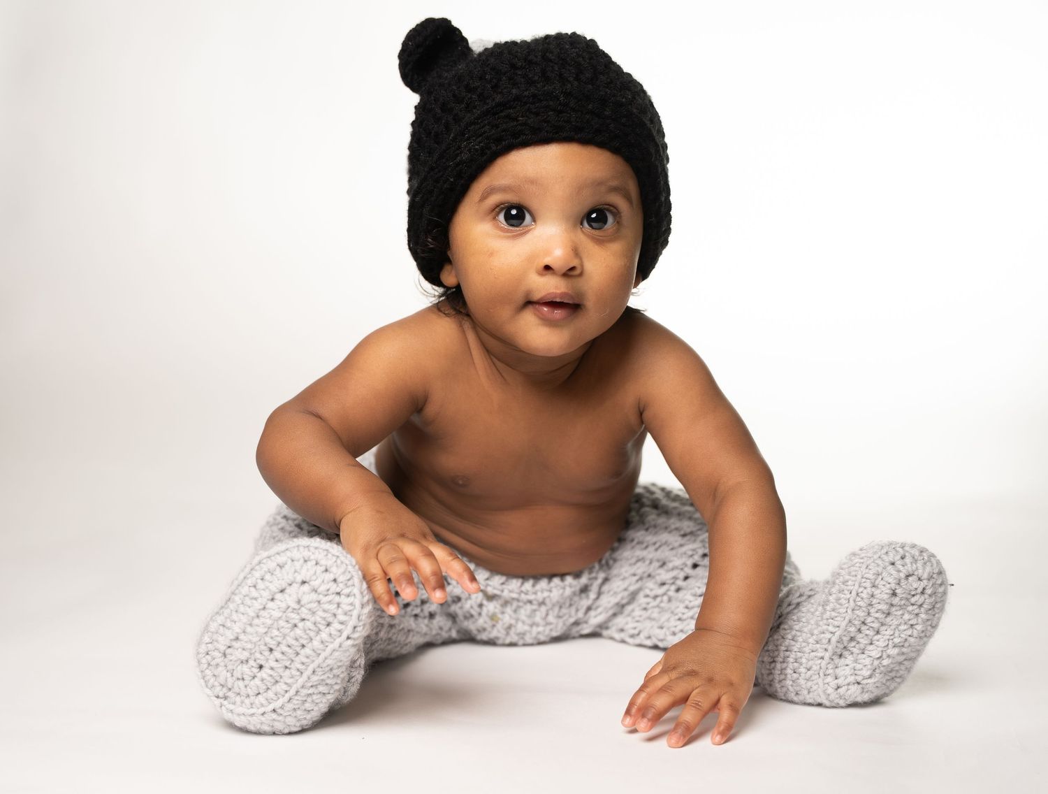 A baby wearing a black hat lies on a white blanket with knit leg warmers in a studio setting.