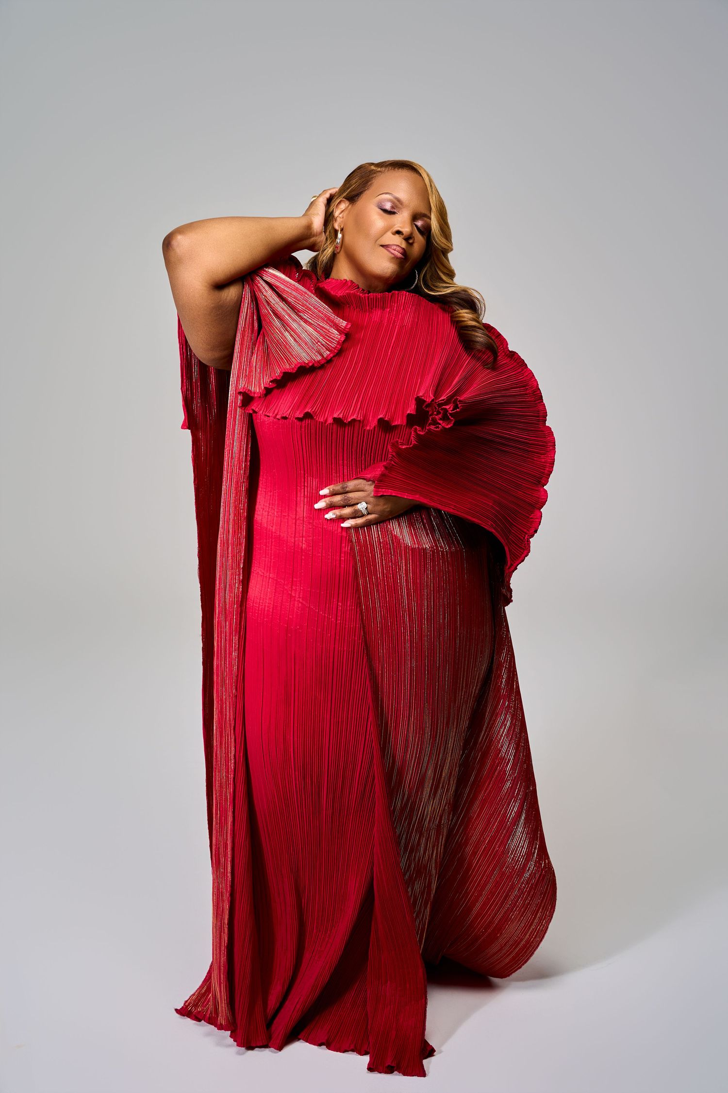 A figure in a flowing red pleated dress poses dramatically against a light gray studio backdrop.