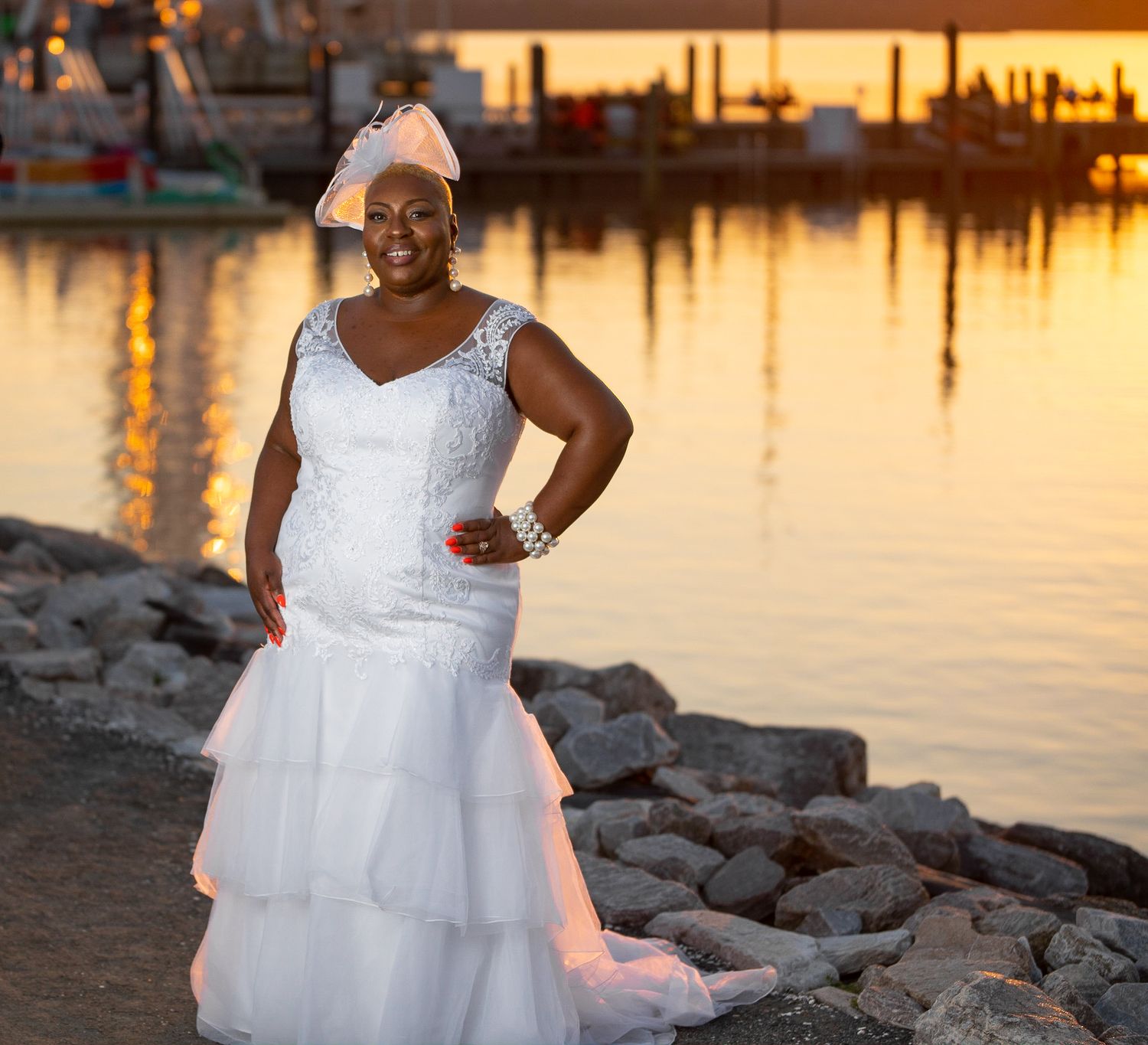 Bride in flowing white dress poses at sunset by a rocky waterfront during golden hour.